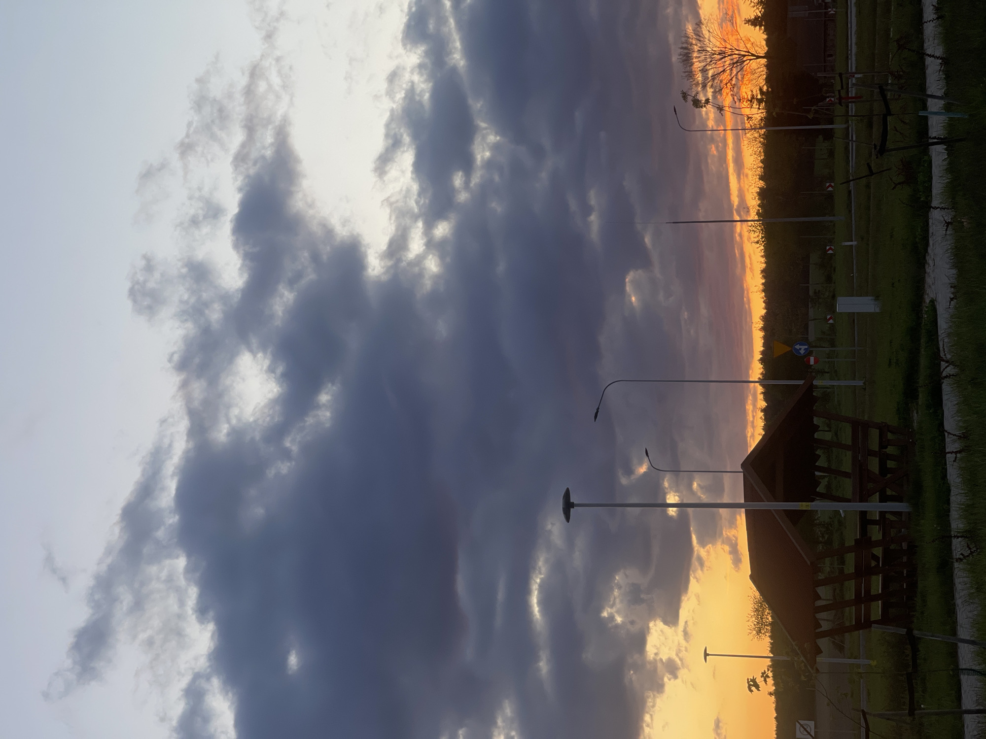 The image depicts a sunset view in Michalinowo, Poland. A large, dark cloud formation dominates the sky on the left, contrasted by the setting sun on the right. The sun casts an orange glow over the horizon, silhouetting trees and vegetation. Several streetlights are visible, as is a wooden structure with a peaked roof. A grassy area and some signage are also present. The scene appears still and quiet.