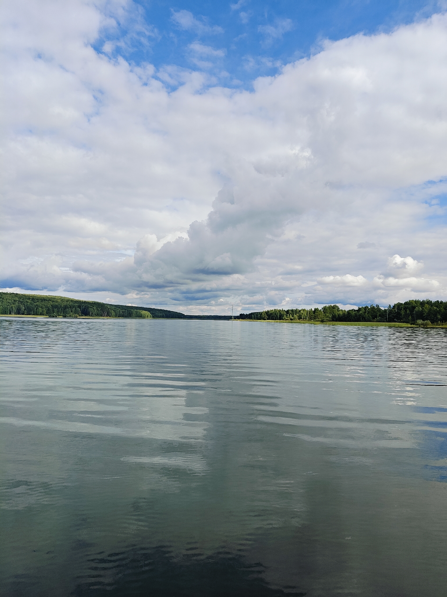 The image depicts a wide, calm body of water, likely a reservoir or river, with a forested shoreline visible in the distance. The sky is filled with a mix of blue sky and white, puffy clouds, with some darker, more ominous clouds on the horizon. The water reflects the sky and surrounding landscape, creating a shimmering effect. The shoreline is densely covered with green trees, indicating a natural, undeveloped area. In the far distance, a tall, slender structure, possibly a radio tower or lighthouse, is barely visible against the tree line. The scene suggests a tranquil, natural environment, potentially a popular spot for outdoor recreation. The location is specified as being near the city of Chaykovsky, Russia, known for its Kama River reservoir, which aligns with the visual characteristics of the image. There are no people or specific actions visible in the frame, making it a purely landscape-focused photograph.