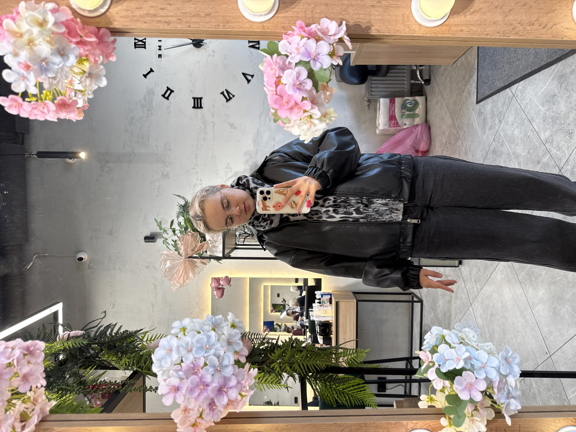 The image shows a woman taking a selfie in front of a mirror. The mirror is framed with wooden panels and decorative artificial flowers. Behind the woman in the mirror's reflection is what appears to be the interior of a commercial space or shop. The mirror's reflection shows a room with a clock, additional mirrors, shelving, and other interior furnishings. The woman is wearing glasses, a black jacket, and dark jeans. She is holding a mobile phone with a patterned case. There are no other immediately identifiable people or significant actions discernible in the image. The environment seems to be indoors. The presence of Roman numeral numbers on the clock visible in the mirror might suggest the location is Saint Petersburg, Russia, given the historical use of Roman numerals in the city's architecture and design. However, this is speculative based on a single element in the image.