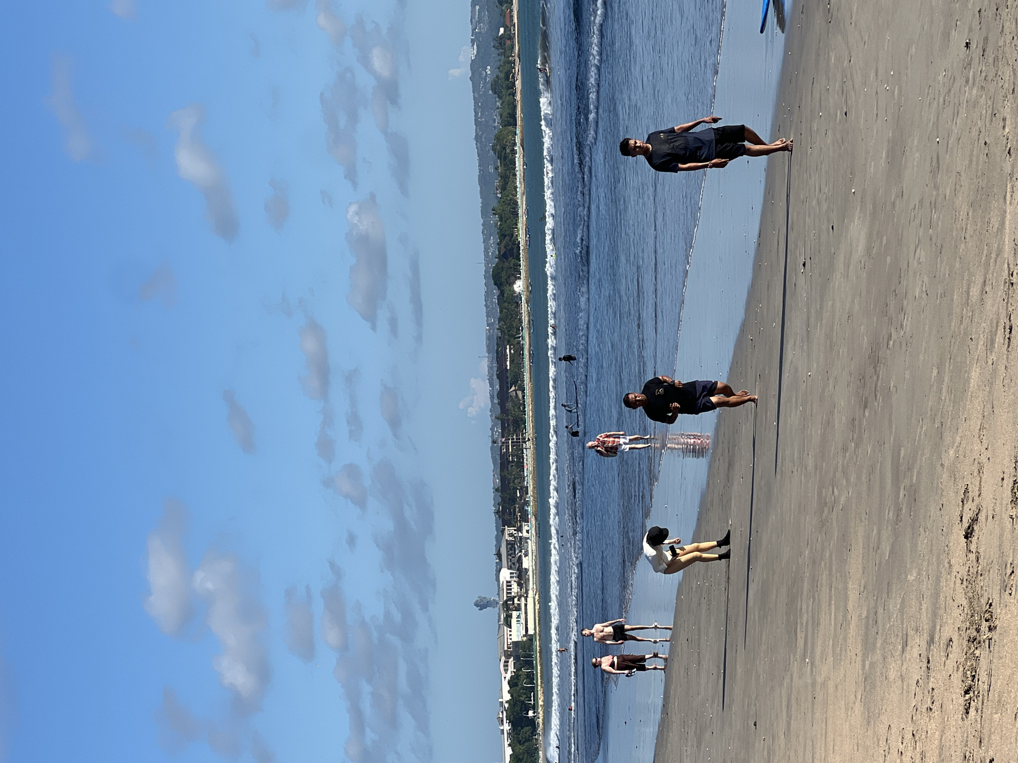 The image depicts a sunny beach scene in Kuta, Indonesia. Several people are visible along the shoreline and in the water. There are two distinct individuals at the top of the frame walking on the beach, with others scattered further down the beach, some appearing to be wading in the water. A clear blue sky with scattered cumulus clouds is visible above. The shoreline is lined with buildings and greenery, indicative of the coastal city setting. Waves are breaking along the beach. The beach appears to be sandy with patches of wet sand closer to the water's edge.