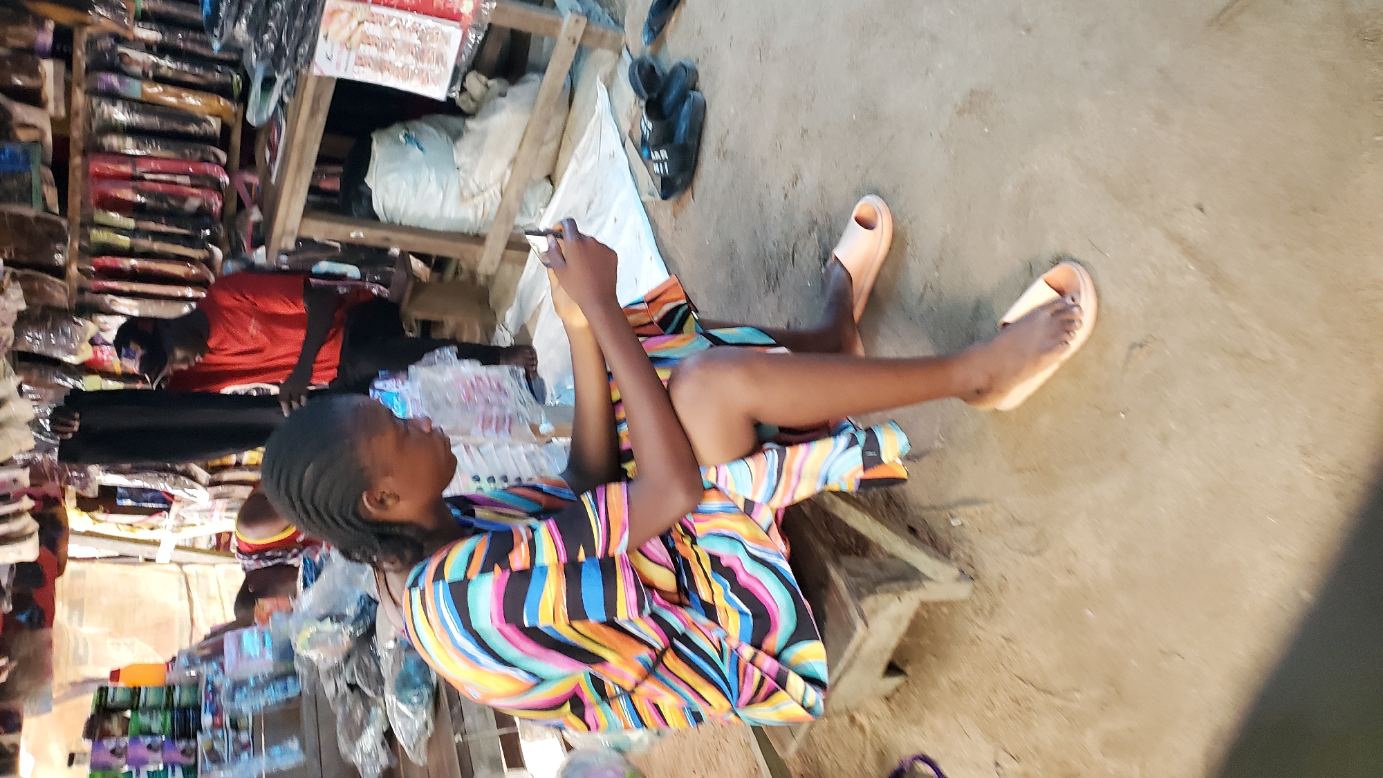 In Jalingo, Nigeria, a young woman wearing a vibrant, multicolored, striped dress sits on a low wooden stool in an outdoor market stall. She is engrossed in her mobile phone, holding it in her right hand. Her left hand rests on her lap. She wears light peach-colored slide sandals on her feet. To her right, on the dusty ground, are two pairs of black slide sandals. In the background, various goods are displayed, including what appears to be hair extensions hanging from racks and products on shelves. A man in a red t-shirt and black cap stands behind a wooden counter, holding up a pair of black trousers. The environment suggests a busy marketplace with a mix of retail and informal commerce.