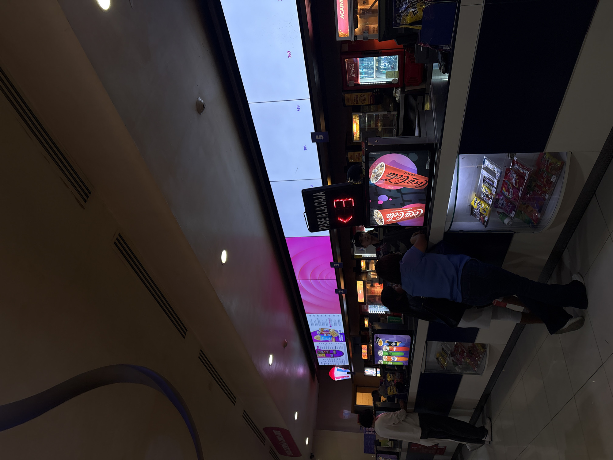 Here's a detailed description of the image:
**General Description:** The image shows the interior of a concession stand at a movie theater. The scene includes digital menu boards, a display of snacks and drinks, and customers being served.
**People:** There are several people visible: customers standing at the counter, possibly ordering or waiting for their food. At least one employee can be seen behind the counter.
**Objects:**
* Digital Menu Boards: Large screens display the menu and prices for items like Coca-Cola, nachos, candy, and other snacks.
* Coca-Cola Refrigerator: A branded Coca-Cola refrigerator is visible, displaying bottles.
* Snacks: Candy, chips, and other snacks are arranged in display cases.
* "Pase a la Caja" Sign: A digital sign directing customers to the checkout counter, with an arrow pointing to the counter.
**Actions/Interactions:** Customers are interacting with the concession stand, presumably ordering and purchasing food and drinks. The employees are likely serving the customers.
**Environment:**
* Lighting: The area is well-lit with overhead lighting.
* Architecture: The ceiling has a curved design.
**Location (Based on OCR and likely context):** The image likely shows the concession stand area of a movie theater in the city of Puebla, Mexico. The brand names and Spanish phrases like "Pase a la Caja" and "Dulceria Caline" point to a location where Spanish is spoken.