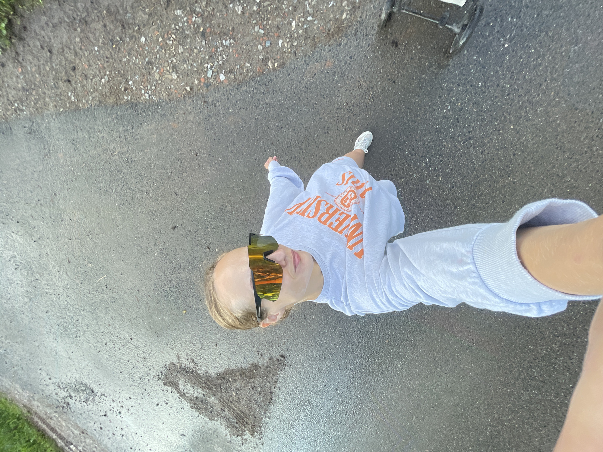 A young woman with blonde hair tied back is taking a selfie outdoors on a wet asphalt surface. She is wearing a light blue sweatshirt with orange lettering that appears to say "UNIVERSITY" and large, reflective sunglasses. Her right arm is extended, holding the phone to take the picture. Her left arm is bent at the elbow. She is wearing white sneakers. The asphalt is dark and appears damp with some water pooling. To the left of the woman, there is a dark, wet patch on the ground. To the right, the asphalt transitions to a gravelly area with scattered small rocks and debris. The overall environment suggests an outdoor, possibly urban or suburban, setting. The image does not contain any specific visual cues to place the location in Подшивалово, Russia.