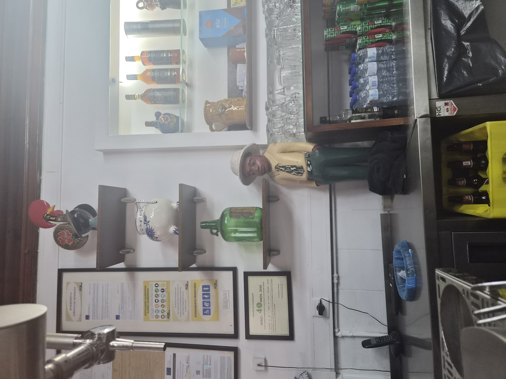 A medium shot captures a dimly lit interior, possibly a bar or restaurant in Barcelos, Portugal, showcasing shelves laden with various items. On the left, a framed document with Portuguese text and a logo is mounted on a white tiled wall, adjacent to a metal beer tap. Above, a decorative ceramic rooster stands sentinel on a wooden shelf. Below, a large green glass bottle is displayed on another shelf, next to a shelf holding a small blue and white ceramic pot.
Dominating the center of the image is a life-sized wooden figurine of a Black man wearing a hat, yellow shirt, and green pants, his hands in his pockets. To his right, a shelf displays a collection of drinking glasses and mugs, with a blue box labeled "DECAF" in the background. Further down, more shelves hold rows of bottled water and canned beverages, including what appear to be beer cans. In the foreground, a stainless steel counter contains a black telephone and a blue plastic bag. The overall impression is one of a functional, perhaps rustic, establishment.