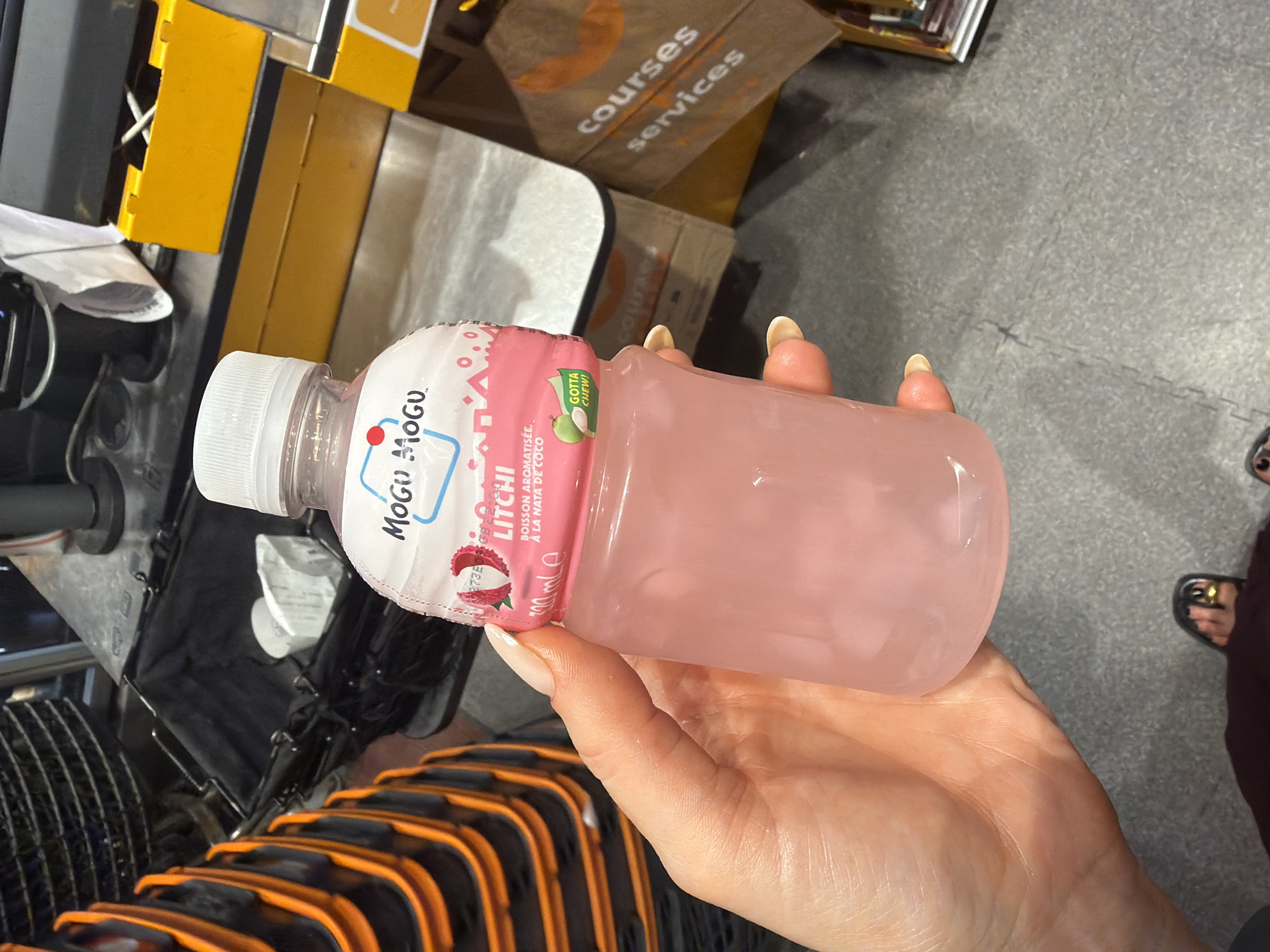 Here is a detailed description of the image:
A person is holding a bottle of Mogu Mogu Litchi drink in their hand. The bottle is pink and partially filled with liquid. The label indicates the drink is flavored with Nata de Coco. The person is in a store near the checkout, with a shopping cart partially visible in the foreground. Behind the drink is a shopping service bag. The location appears to be in a grocery store in Paris, France.