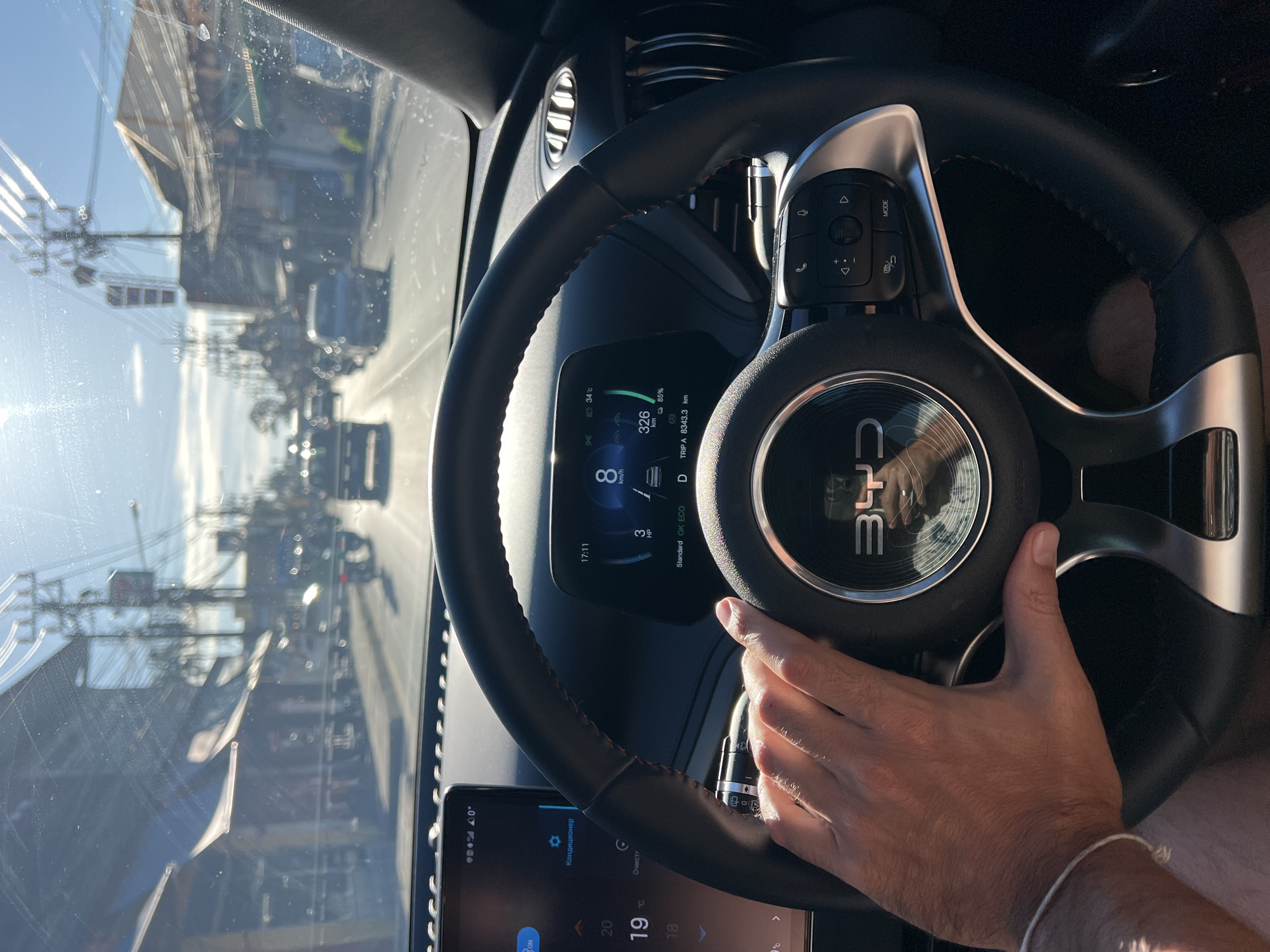 The image shows a first-person perspective from inside a vehicle driving down a street in Baan Mae Nam, Thailand. The road is lined with buildings and overhead power lines. The sun is shining brightly, creating glare on the windshield. The dashboard and steering wheel of the car are visible, with the driver's hand resting on the steering wheel. The car's digital display shows a speed of 8 km/h and a temperature of 34°C. Other vehicles are present on the road, including cars and motorcycles. The scene depicts a typical day on a street in a Thai town.
