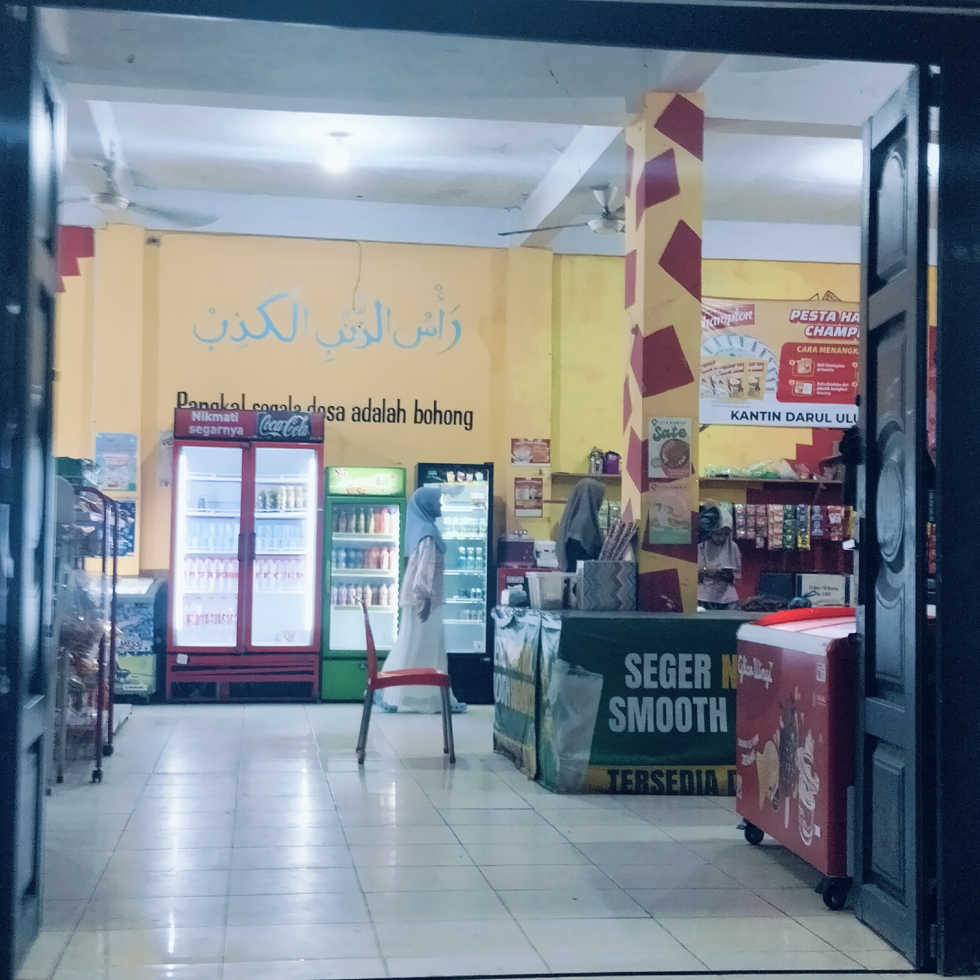 This image captures the interior of a small shop or canteen, likely located within an educational or religious institution, given the prominent text "KANTIN DARUL ULU." The setting features bright yellow walls with a red and yellow checkered pillar, and a white tiled floor, giving it a clean yet utilitarian appearance.
Two individuals, both appearing to be women wearing hijabs, are visible within the space. One stands near a green refrigerator, facing away from the camera, dressed in a light-colored garment. The other is partially obscured behind a counter on the right side of the frame, suggesting they might be attending to customers or working.
The shop is well-stocked with various items. Several upright refrigerators, including a red Coca-Cola branded one, a green one, and a black one, display beverages. A smaller red cooler on wheels is positioned near the entrance on the right. A red plastic chair with metal legs is placed in the middle of the floor. Behind the counter, shelves hold products, and a banner advertises "PESTA HA CHAMP" with "CARA MENANGK" (How to Win). Another banner on the counter reads "SEGER SMOOTH TERSEDIA" (Fresh Smooth Available).
Notable text on the yellow wall includes Arabic calligraphy "رَأْسُ الرتب الكذب" (Ra'su al-rutab al-kadhb), directly translated below in Indonesian as "Pangkal segala dosa adalah bohong" (The root of all sin is lying). A Coca-Cola advertisement encourages "Nikmati segarnya Coca-Cola" (Enjoy the freshness of Coca-Cola). The overall scene depicts a typical, quiet moment in a canteen, likely during daytime, with a focus on daily commerce and provisions.