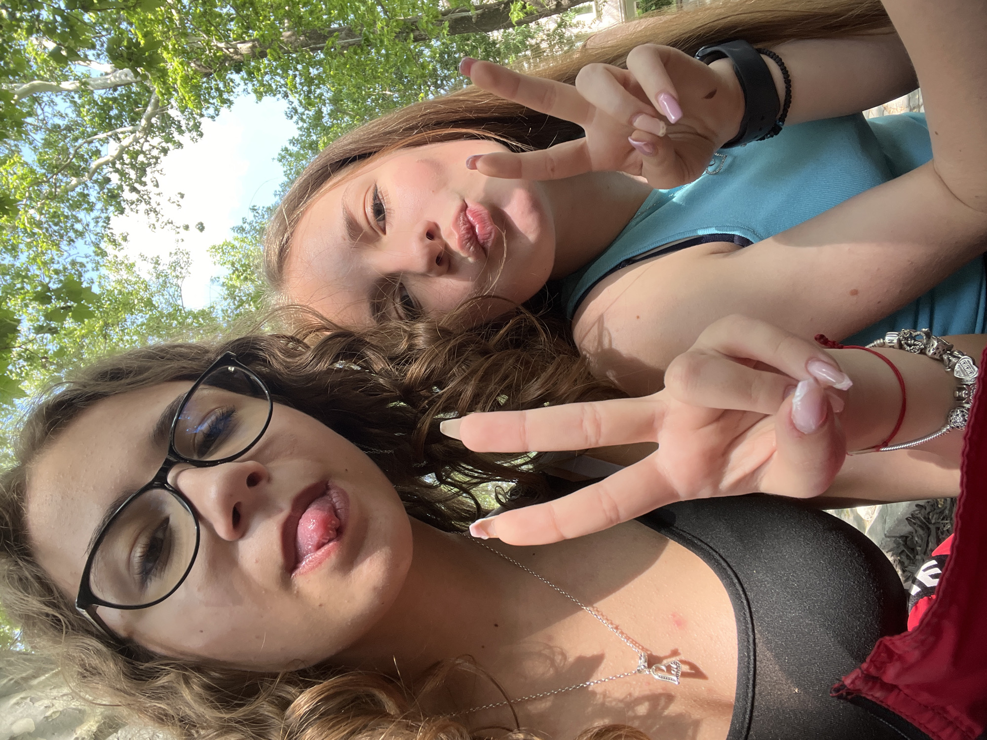Two young women are taking a selfie from a low angle. The woman in the foreground wears glasses, a black top, and a silver heart necklace, and sticks her tongue out. The woman in the background wears a blue top and makes a kissing face. Both women are making a "peace" sign with their fingers. The background shows green trees and a building, indicating they are outdoors. Given the location constraints, a possible location for the image is in a park in Plovdiv, Bulgaria.