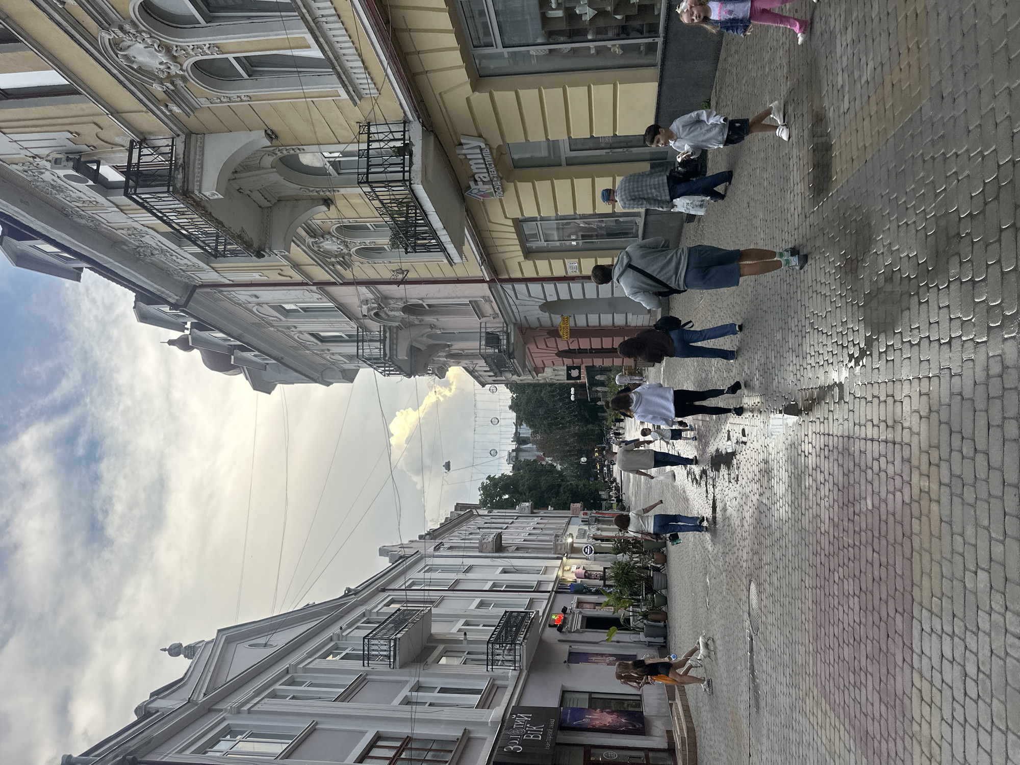The image captures a street scene in Ternopil, Ukraine. In the foreground, a paved street with some wet patches suggests recent rain. Several people are walking along the street. On the left, a woman and a girl walk with a small white dog on a leash. Further down, a group of people are walking away from the camera, some carrying shopping bags. On the right, a young boy in a grey hoodie and shorts walks with a child in a pink outfit and a denim jacket. The street is lined with buildings that feature traditional European architecture, with balconies and ornate facades. The sky is overcast with some dramatic cloud formations. Several shops are visible along the street, indicated by signs and window displays. Decorative lights or lanterns are strung across the street in the distance, adding to the atmosphere of a city center. The overall impression is of a bustling urban environment on a possibly damp day.