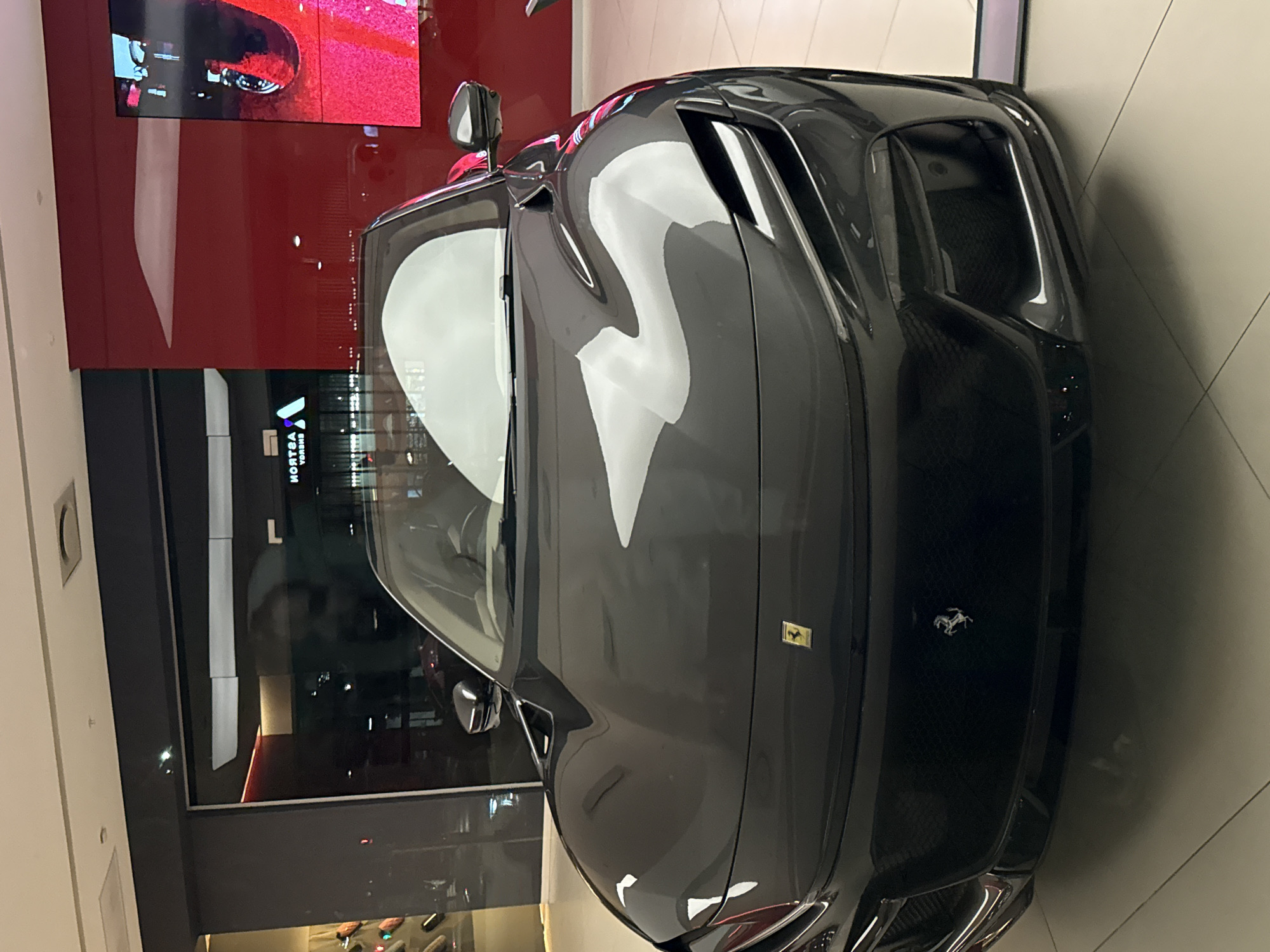 A dark grey Ferrari Purosangue is centrally positioned on a white tiled floor within a brightly illuminated luxury car showroom, likely located at the V&A Waterfront in Cape Town, South Africa. The vehicle faces the viewer, displaying its front grille with a silver prancing horse emblem and a yellow Ferrari shield badge on the hood. The car's polished surface reflects overhead lighting and elements of the showroom. Behind the vehicle, a large glass storefront reveals reflections of interior ceiling structures and reversed text, including "ASTROA ENERGY." To the right, a prominent red wall features an abstract white design and a large digital screen displaying a vivid red and black image. Dark grey structural elements frame the glass partitions, and recessed lighting is visible on the ceiling. No individuals are depicted in the scene.