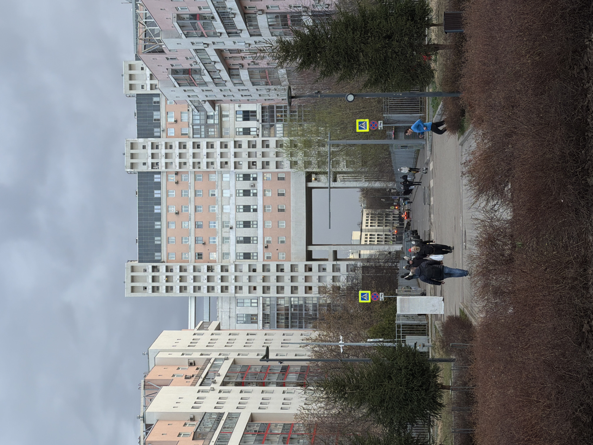 The image shows a view of a street in Khimki, Russia. Several high-rise apartment buildings are visible in the background, constructed of concrete and painted in pastel colors. The sky is overcast. A sidewalk is visible in the foreground, lined with bare bushes and trees. Several people are walking along the sidewalk. Two street signs are visible, one with a pedestrian crossing symbol and the other with a 'no parking' symbol.