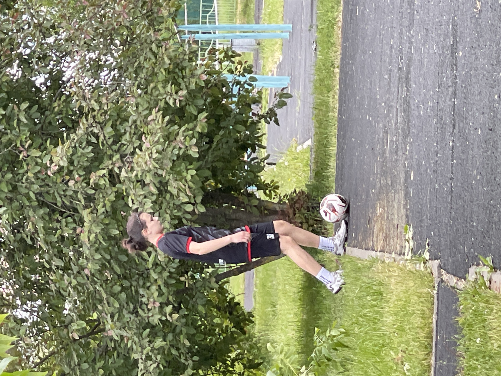 A man with long hair tied back is outside in a grassy area next to a paved path. He is wearing a black and red sports jersey, black shorts, white socks, and white athletic shoes. A soccer ball rests on the edge of the paved path next to the man's foot. A leafy tree is situated to the left of the man, and a blue metal fence can be seen in the background. The location is likely in the city of Moscow, Russia.