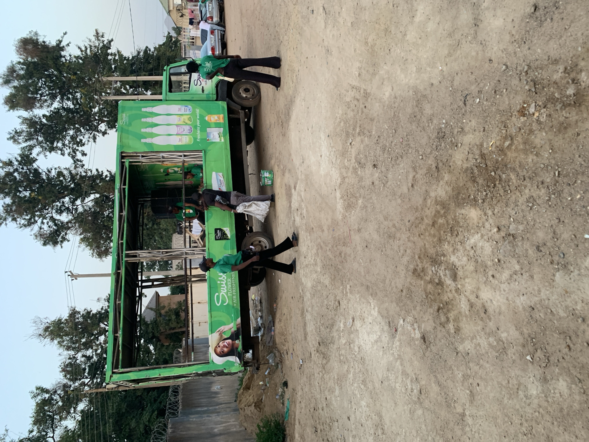 The scene depicts a green Swiss Flower air freshener promotional truck in an open, unpaved area near New Stadium, Nigeria. Two individuals wearing green shirts and black pants are positioned near the truck, with one possibly loading or unloading materials. Litter and debris are scattered on the ground. In the background, trees, a barbed wire fence, and other vehicles are visible. The sky is overcast.