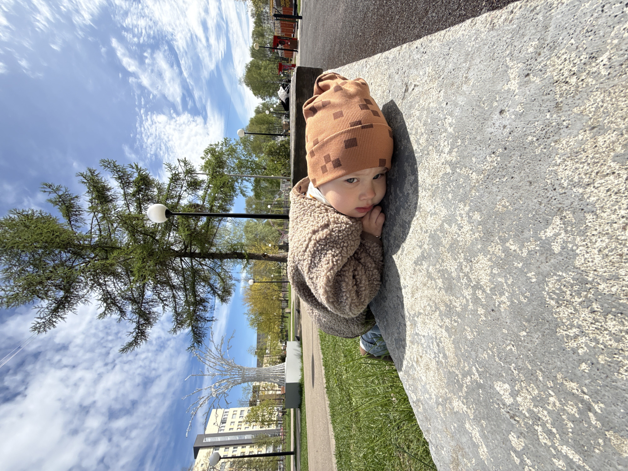 A young child with a brown and tan checkered hat and a tan jacket sits with their face resting on their hand near a gray concrete bench in a city park in Unknown, Unknown. The child is positioned to the right of the frame. Behind the child is a park, with a green lawn, trees, street lamps, and a white decorative metal tree sculpture. A building is visible in the background. The sky is blue with scattered white clouds.