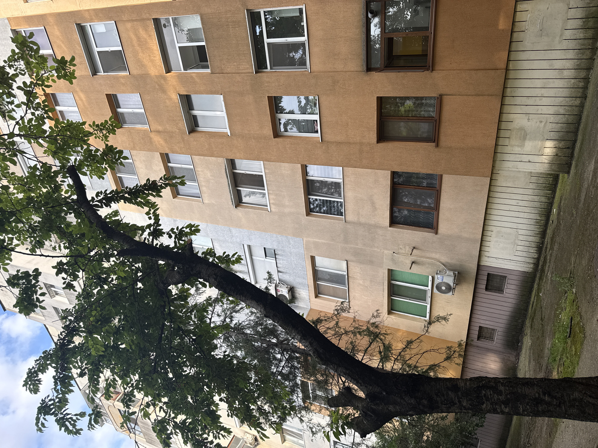 The image depicts a view of an apartment building with a large tree in the foreground, obscuring a portion of the structure. The building appears to be in a city, likely Stara Zagora, Bulgaria, given the architectural style and the presence of air conditioning units visible on the exterior walls. The building's facade is painted in varying shades of beige and orange, with multiple windows of different sizes and configurations. Some windows are open, while others are closed with blinds or curtains. The tree in the foreground has a thick, dark trunk and branches covered in green leaves, suggesting it is a deciduous tree during its growing season. The ground area in front of the building appears to be a paved walkway or courtyard with some patches of grass and dirt. The sky above is partly cloudy. The overall scene is that of an urban residential area.