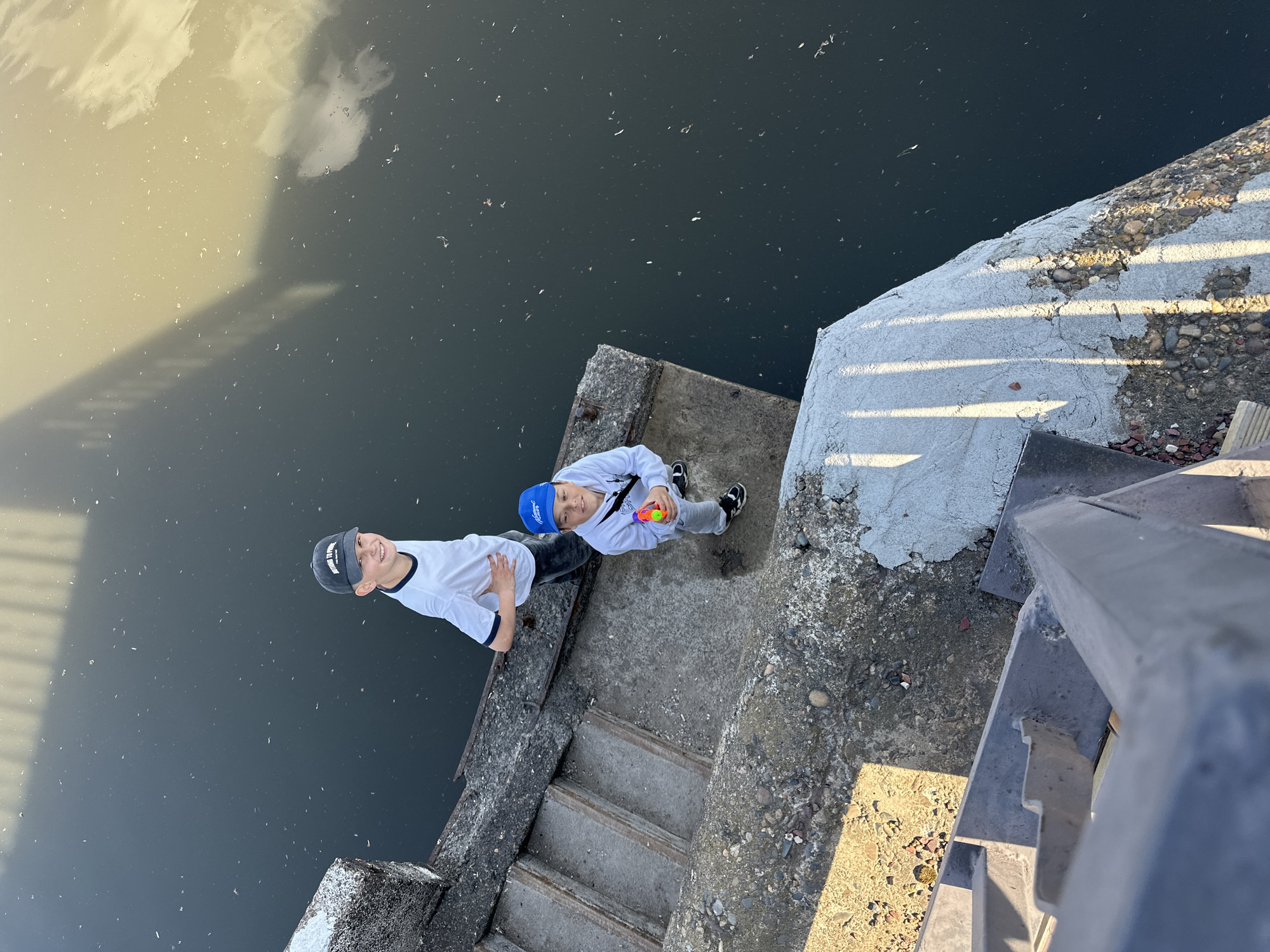 Two young boys stand on a concrete platform overlooking a body of water, presumably the Kama River in Chaikovsky, Russia. The boy on the left wears a white long-sleeved shirt with dark trim, a dark baseball cap, and dark pants. The boy on the right wears a white hooded sweatshirt, a blue baseball cap, and grey pants, and holds a toy water gun. They are positioned on a concrete structure with stairs leading down. The body of water appears dark and reflects the sky. A concrete barrier with a metal railing is visible to the right.
