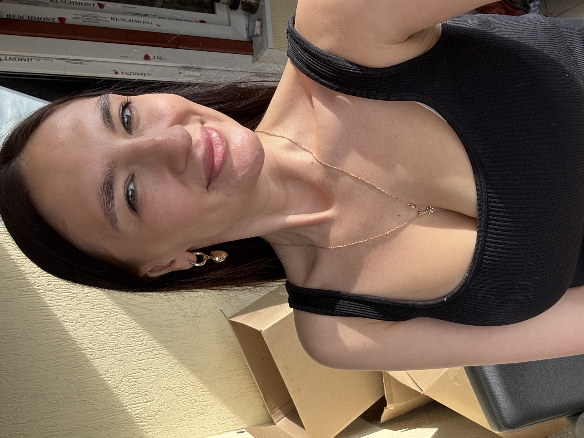 A woman is posing outdoors on a sunny day. She has dark hair and green eyes, and is wearing a black tank top and gold jewelry (necklace and earrings). She is sitting on a black object near cardboard boxes, with a light yellow textured wall in the background. There is a window visible in the top of the image labeled "Reachmont." There is no definitive evidence to pinpoint the location to Sochi, Russia.