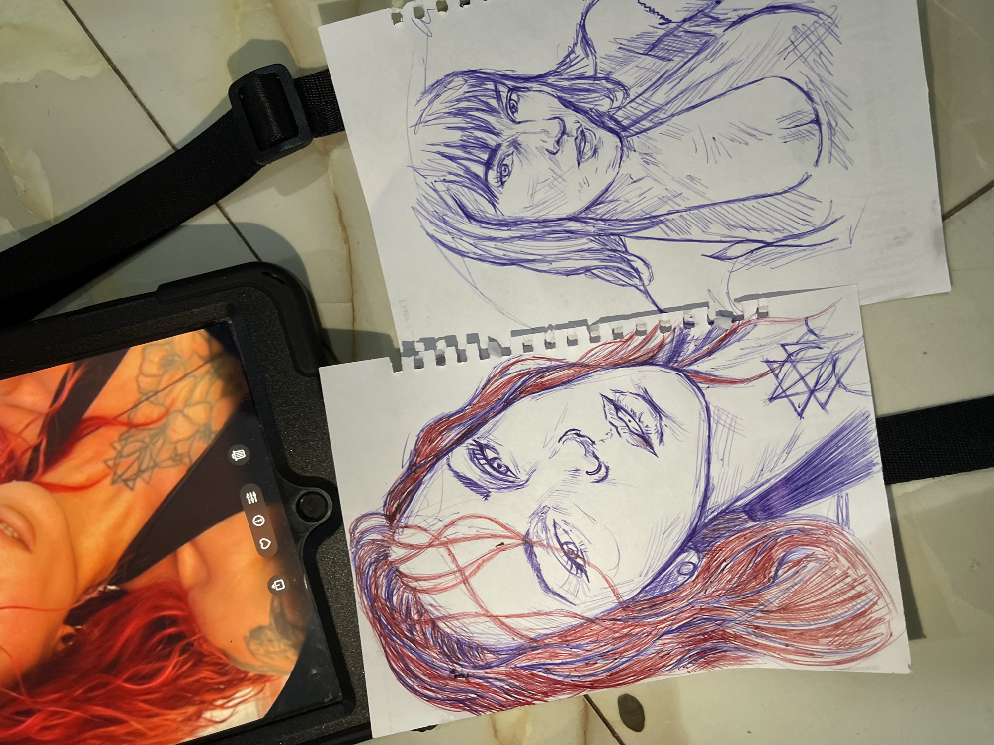 The image shows two pen and ink sketches of women's faces on separate sheets of paper, placed on a tiled floor. The sketch on the left is in red and blue ink, depicting a woman with red hair looking towards the viewer. The sketch on the right is in blue ink, showing a woman with bangs and long hair, also looking towards the viewer. In the background, a tablet displays a close-up image of a woman with red hair and tattoos. A black strap from a bag is visible across the tablet and extends towards the bottom right of the image. The scene appears to be indoors, with a tiled floor visible. The location is not specified in the image.