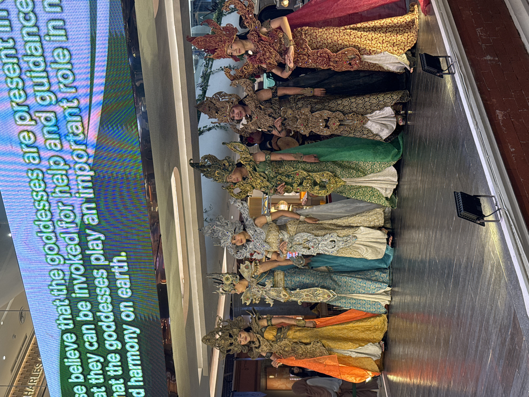 This image captures a cultural event or performance taking place indoors, likely in Denpasar, Indonesia. Eight women are prominently featured, standing in a line on a dark stage or platform. They are all dressed in elaborate, traditional-style costumes that appear inspired by Indonesian cultural attire. Each costume features an ornate headpiece, often golden or silver and highly detailed, a richly decorated top, and a long, flowing skirt in various colors including red, gold, green, white, blue, and orange.
The women are posing, generally looking towards the viewer or slightly off to the side, with serene or neutral expressions. Their attire, particularly the intricate headwear and embellishments, suggests they might be portraying deities or characters of cultural significance, possibly goddesses. The stage is illuminated by several spotlights visible on the floor.
To the left of the image, a large digital screen displays text, providing context for the event. The visible text reads: "believe that the goddesses are present, that they can be invoked for help and guidance, that the goddesses play an important role in and harmony on earth." This suggests the event is themed around spiritual or mythological goddesses. In the background, elements of an indoor venue, possibly a shopping mall, are visible, including a partial "CHARLES & KEITH" sign. The overall scene conveys a rich display of cultural heritage and artistry.