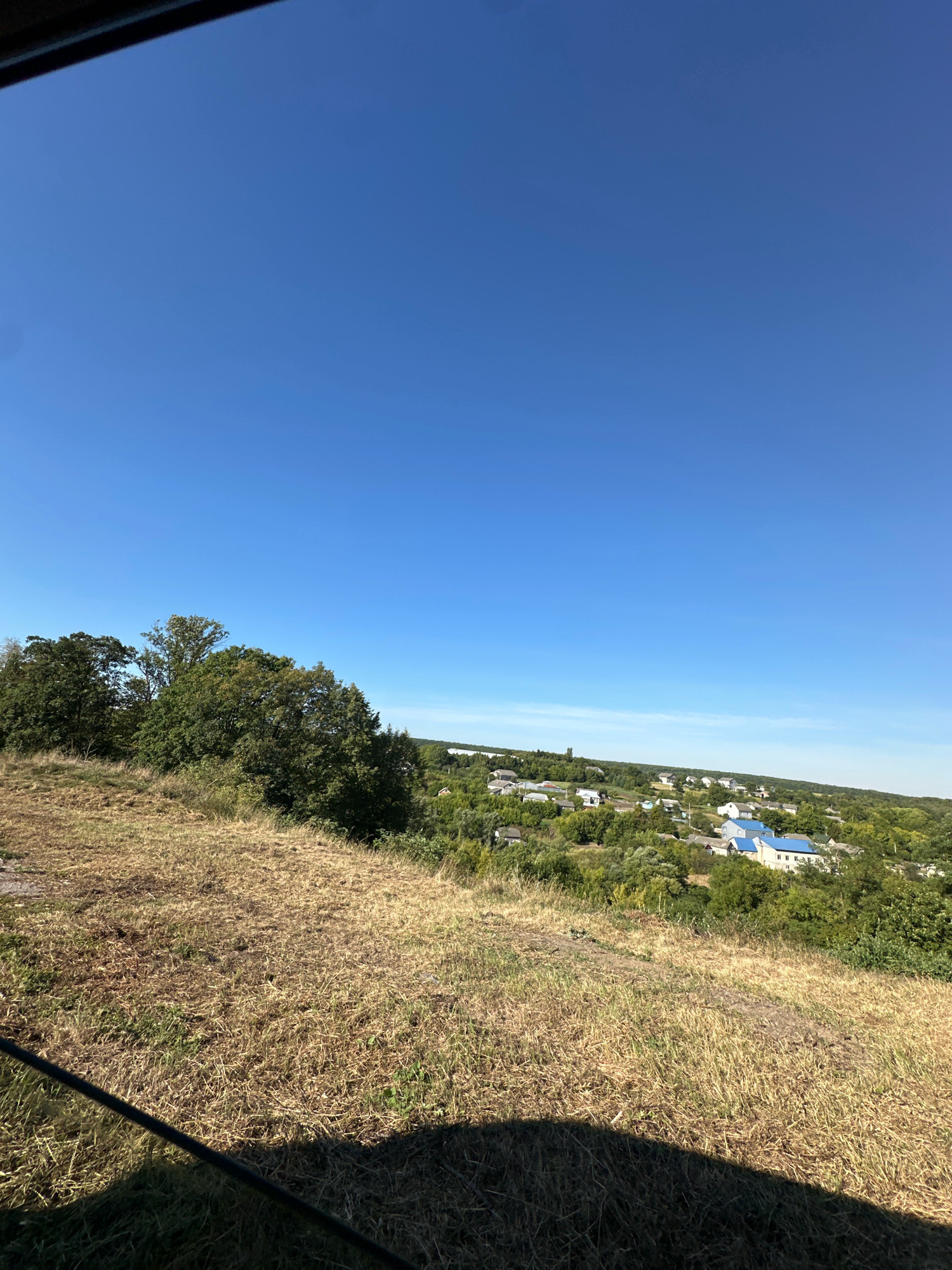 This is a daytime, eye-level, full outdoor shot of a landscape in or around Makiv, Ukraine. The foreground is dominated by a dry, grassy hill, casting a long shadow from the left. In the mid-ground, a dense collection of green trees and bushes is visible. Beyond the trees, a village or town is nestled within the rolling hills, characterized by numerous houses with various roof colors, including prominent blue roofs on several buildings. The sky above is a clear, deep blue.