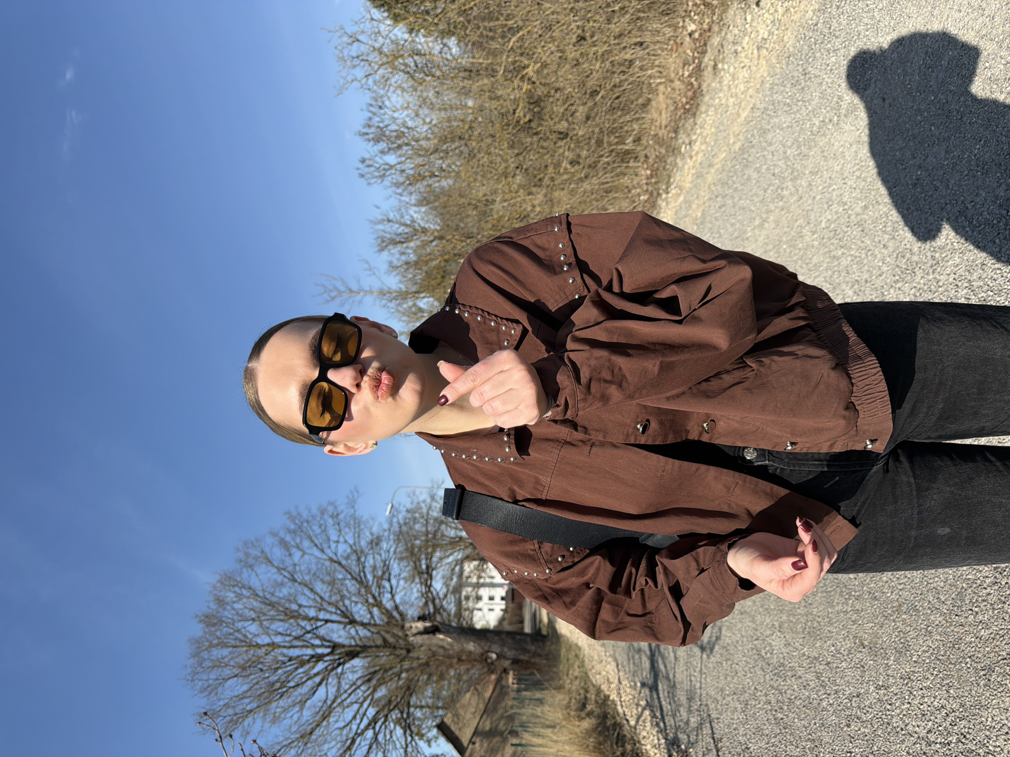 A woman stands on a paved road under a clear blue sky. She is wearing a brown jacket with metal studs along the shoulders and collar, black jeans, and yellow-tinted sunglasses. She is blowing a kiss toward the camera. A black strap crosses her torso, likely part of a bag or camera. Behind her are bare trees and what appears to be a residential building. To the right, a shadow of what might be a person or object is visible on the road. Based on the environment and available information, the location is potentially Dreiliņi, Latvia.