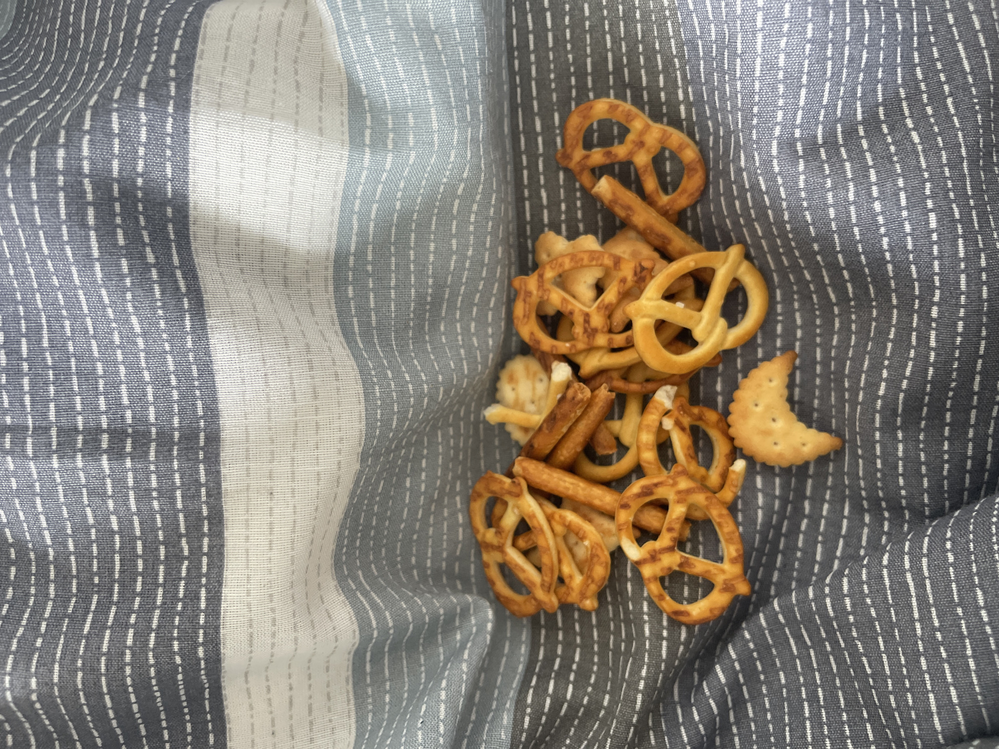 A pile of pretzels and cheese crackers rests on a striped gray and white fabric. The pretzels are mostly standard shapes, and the crackers are crescent-shaped. The location is unknown, and there's no indication of being in Sofia, Bulgaria. No people are present.