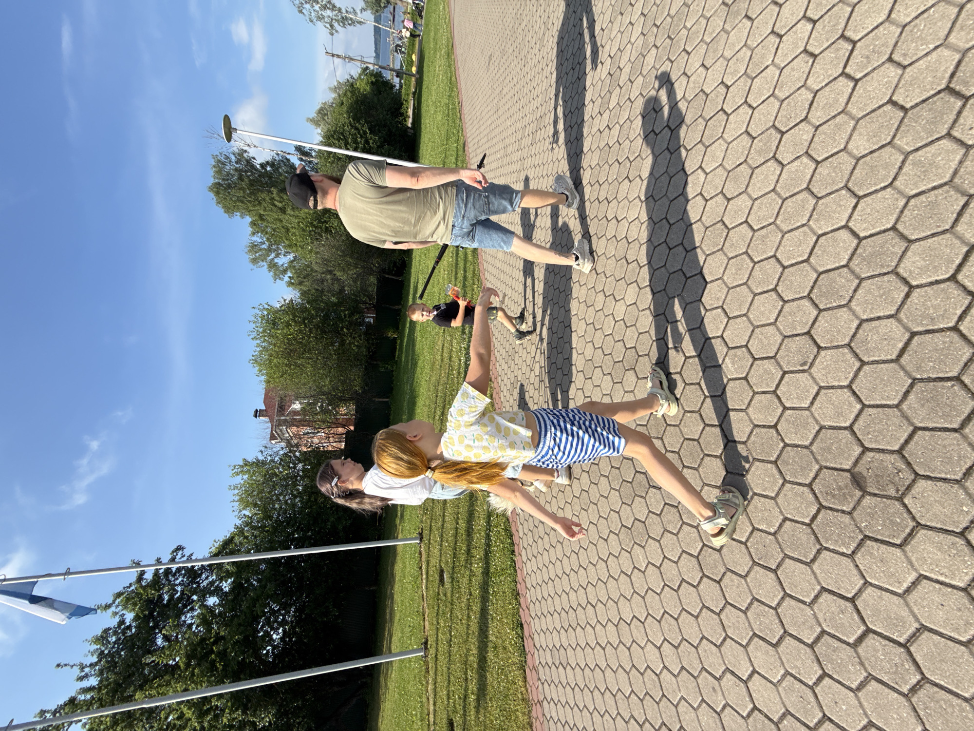 A group of people, including two girls, a young boy, and a man, are walking on a hexagonal-patterned paved walkway in Valday, Russia. The walkway appears to be in a park or public area, with a grassy lawn and trees to the left, and a body of water and buildings visible in the background to the right.
The girl in the foreground, with red hair pulled into a ponytail, is wearing a white crop top with a lemon pattern and blue and white striped shorts. She is walking away from the camera, with her left arm extended. Beside her, slightly behind, a second girl with long brown hair, wearing a white top and jeans, is also walking away from the camera.
A young boy in a black t-shirt and shorts is walking ahead of the man, holding a red object. The man, wearing a green t-shirt and denim shorts, is walking towards the right of the frame, carrying a long, thin object.
A Finnish flag is visible on a pole to the left of the frame, against a clear blue sky with some white clouds. The sun is shining, casting shadows on the walkway. The overall impression is of a casual outdoor outing.