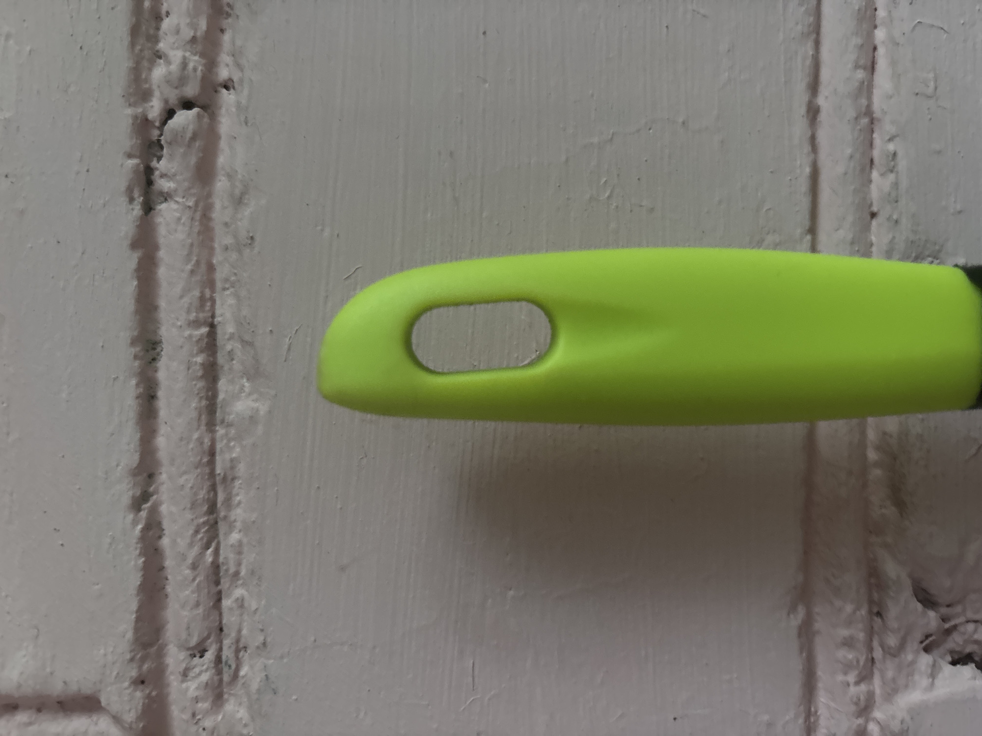 A close-up, eye-level shot captures the top portion of a bright lime green utensil handle against a white brick wall. The handle is smooth and curved, featuring an oval-shaped opening near its top. The brick wall behind it displays visible mortar lines and a textured, slightly weathered surface.