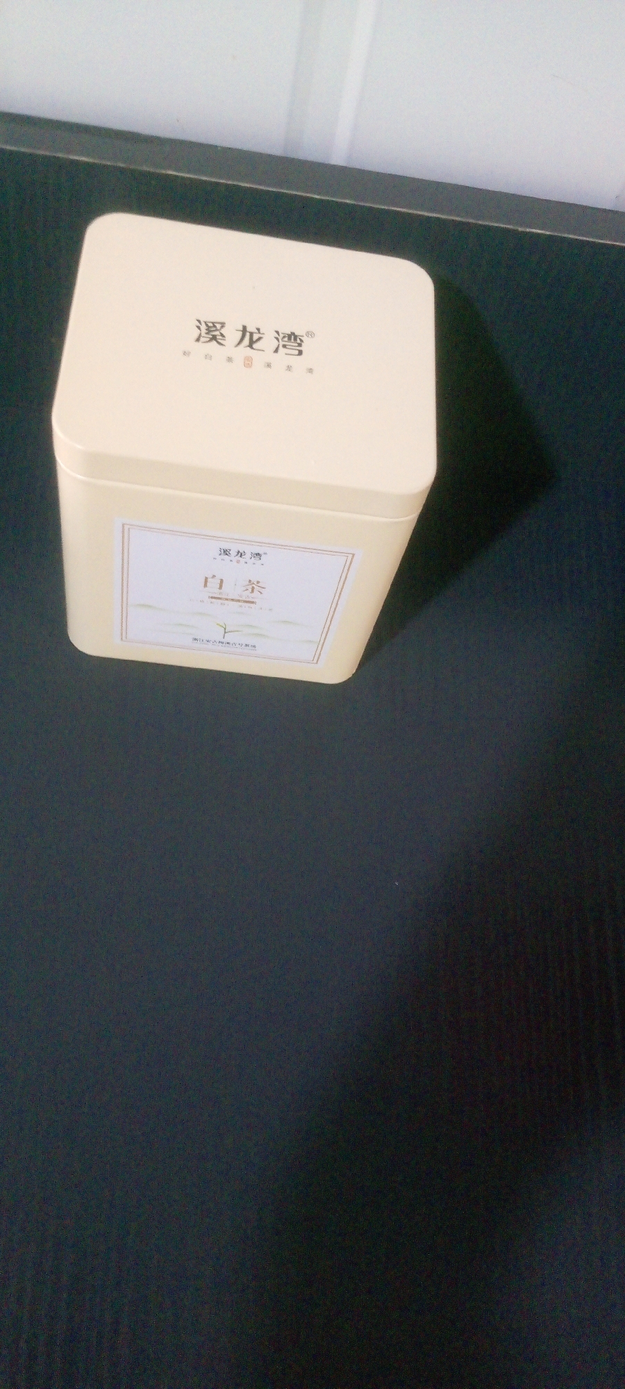 A close-up, high-angle shot shows a light yellow square tin of tea on a dark surface. The lid of the tin displays the brand name "溪龙湾" (Xī Lóng Wān) in Chinese characters. A label on the front of the tin also features the brand name and the words "白茶" (Bái Chá), meaning white tea, in larger characters. Below the tea type, a stylized image of a tea leaf is visible, and at the bottom of the label, there is more Chinese text. The tin appears to be resting on a textured, dark surface, possibly a table or desk. The lighting is somewhat dim, with a shadow cast on the right side of the tin and the surface. The background is out of focus, showing a light-colored wall with vertical lines. The overall scene is simple and focused on the product packaging. The image was taken in Indo, Nigeria.