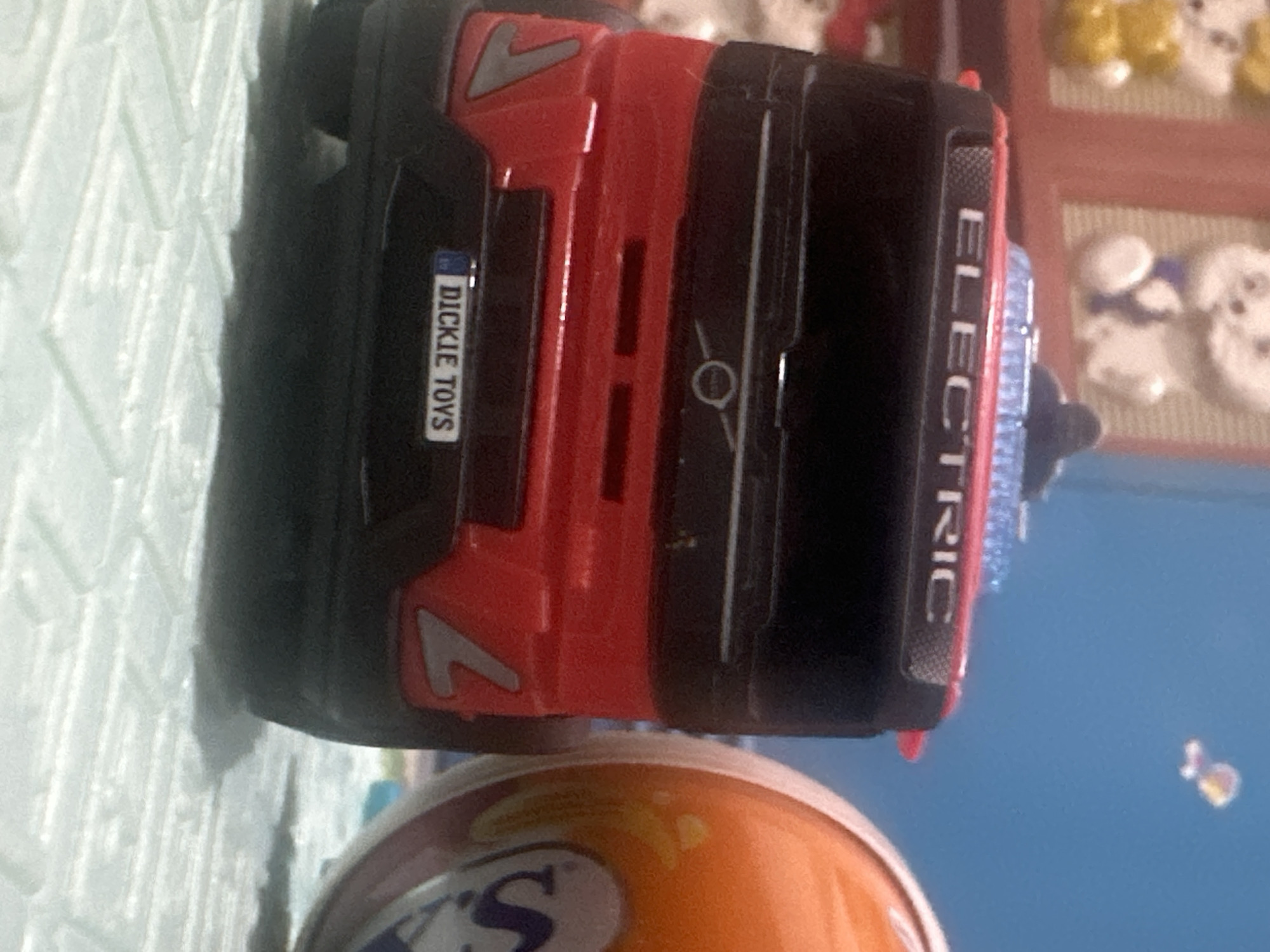 A close-up, low-angle shot shows the rear of a red and black toy truck. The truck has "ELECTRIC" written in white letters across the top. Below that, the rear bumper displays a license plate that reads "DICKIE TOYS." To the right of the truck, a portion of a round, orange and white object is visible, possibly a toy. The background is blurry, showing a light blue wall with a small, indistinct object in the upper right corner. The foreground features a textured, light-colored surface, likely a mat. The scene is indoors, with no clear indication of the City of Binjai, Indonesia.