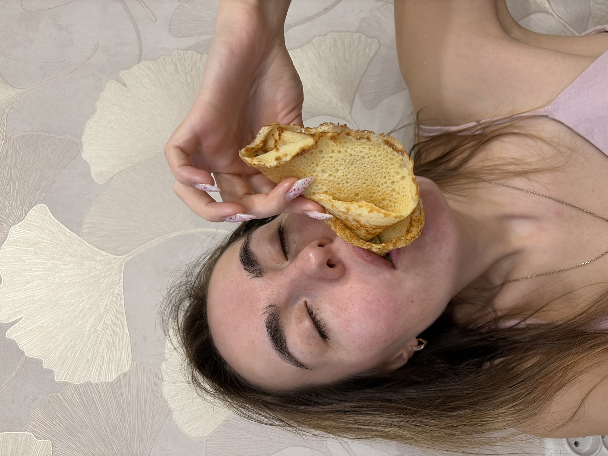A woman with closed eyes is holding a folded pancake towards her open mouth. She has light skin, brown hair, and is wearing a light pink top. The background features a wallpaper with a ginkgo leaf pattern in shades of beige and grey. The overall scene suggests it is indoors. There is no visible information to suggest a location in the city of Izhevsk, Russia.