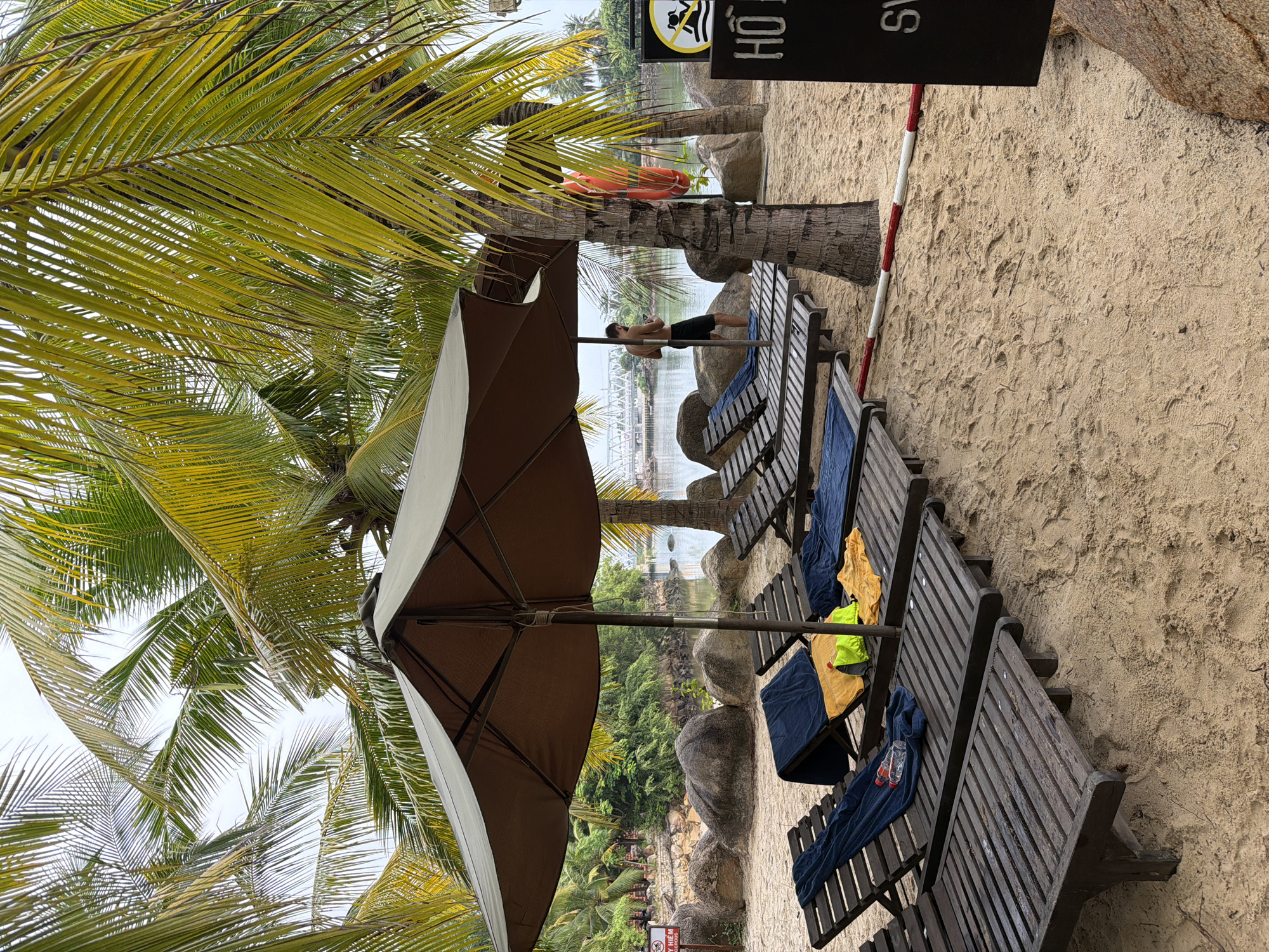 A sandy beach in Nha Trang, Vietnam, is depicted with rows of empty wooden lounge chairs. These chairs are draped with various towels and a few water bottles. A large brown umbrella provides shade over some of the chairs. In the background, lush palm trees frame the scene, with a body of water visible beyond the beach. A man is standing near the water, facing away from the camera, partially obscured by a palm tree. There is a red life preserver attached to a pole. A black sign with white text, partially visible, is positioned on the right side of the image. The sky is overcast, suggesting a cloudy day.