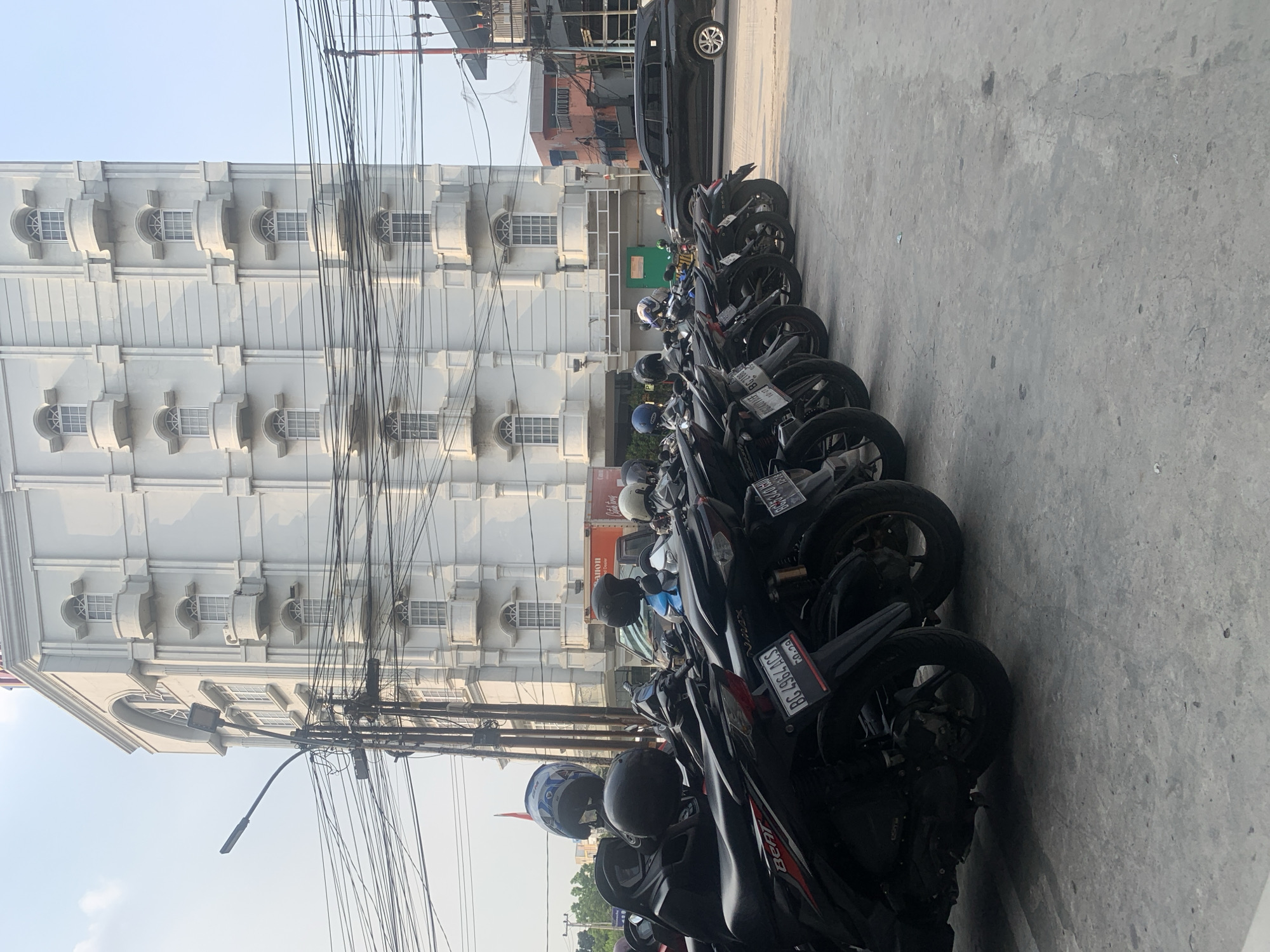 The image shows a row of parked motorcycles on a concrete surface in front of a tall, white building with a classical architectural style. The building has multiple stories, each with a series of arched windows and small balconies. Overhead, a complex network of power lines crisscrosses the sky. A black SUV is visible on the road in the background, near a smaller orange building. The scene appears to be outdoors during the daytime, with a clear sky. The license plate of one of the motorcycles is partially visible, reading "BG 4964 AGS". This indicates the image was likely taken in Palembang, Indonesia, as "BG" is the vehicle registration code for South Sumatra, the province where Palembang is located.