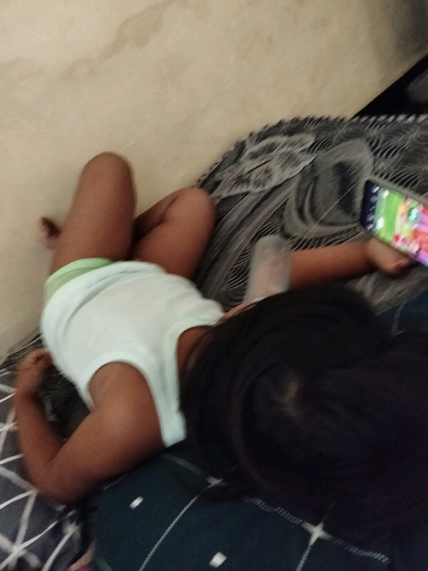 The image shows a child, likely a toddler, lying on a bed. The child is wearing a white tank top and a light green diaper. Their legs are bent, and their left arm is resting on their stomach, while their right arm is extended. The child's head is turned to the right, and their dark hair is visible. The child is holding a smartphone in their right hand, which is displaying a colorful game.
The background consists of a bed with dark patterned bedding and a light-colored wall or surface behind it. The scene appears to be indoors, in a casual setting, possibly a bedroom. The lighting suggests it could be daytime, though the exact time is not clear. The overall mood seems relaxed, with the child engrossed in their game. The location is identified as Gempolan, Indonesia. There is no visible text in the image.