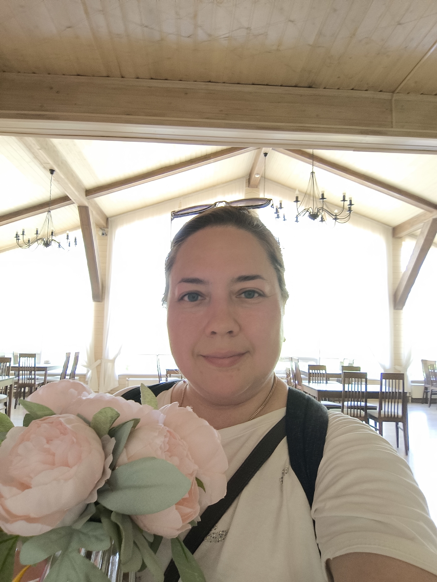 A woman with light skin and brown hair pulled back is taking a selfie indoors, holding a bouquet of light pink peonies. She is wearing a white t-shirt and a dark backpack strap is visible across her shoulder. She has a small necklace on. Behind her, the interior of a room with a high, wooden beamed ceiling is visible. There are tables and chairs, suggesting a restaurant or dining area. Large windows with sheer curtains are in the background. The lighting is bright, suggesting daytime. The image does not contain any specific landmarks or contextual clues to indicate the city of Taldom, Russia.
