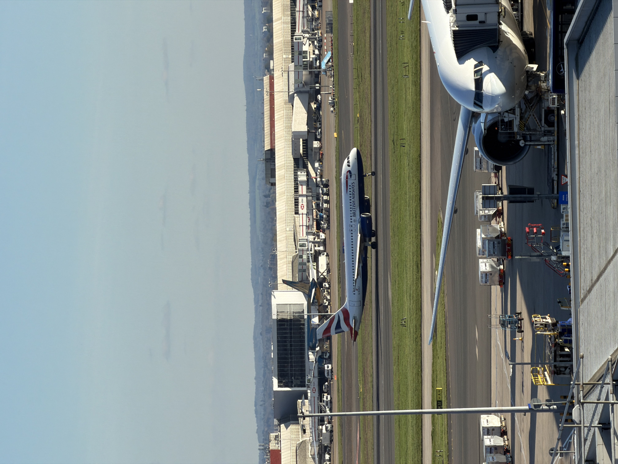 The image shows an aerial view of an airport, likely Heathrow Airport in London, United Kingdom, due to the presence of British Airways airplanes. The sky is clear. In the foreground, there is a section of a building with scaffolding. Adjacent to it is an airplane, docked with a jet bridge. In the middle ground, another British Airways plane is seen taxiing on the runway, alongside the terminal buildings. Various airport vehicles and equipment are visible on the tarmac. In the background, a distant cityscape and hills can be seen.