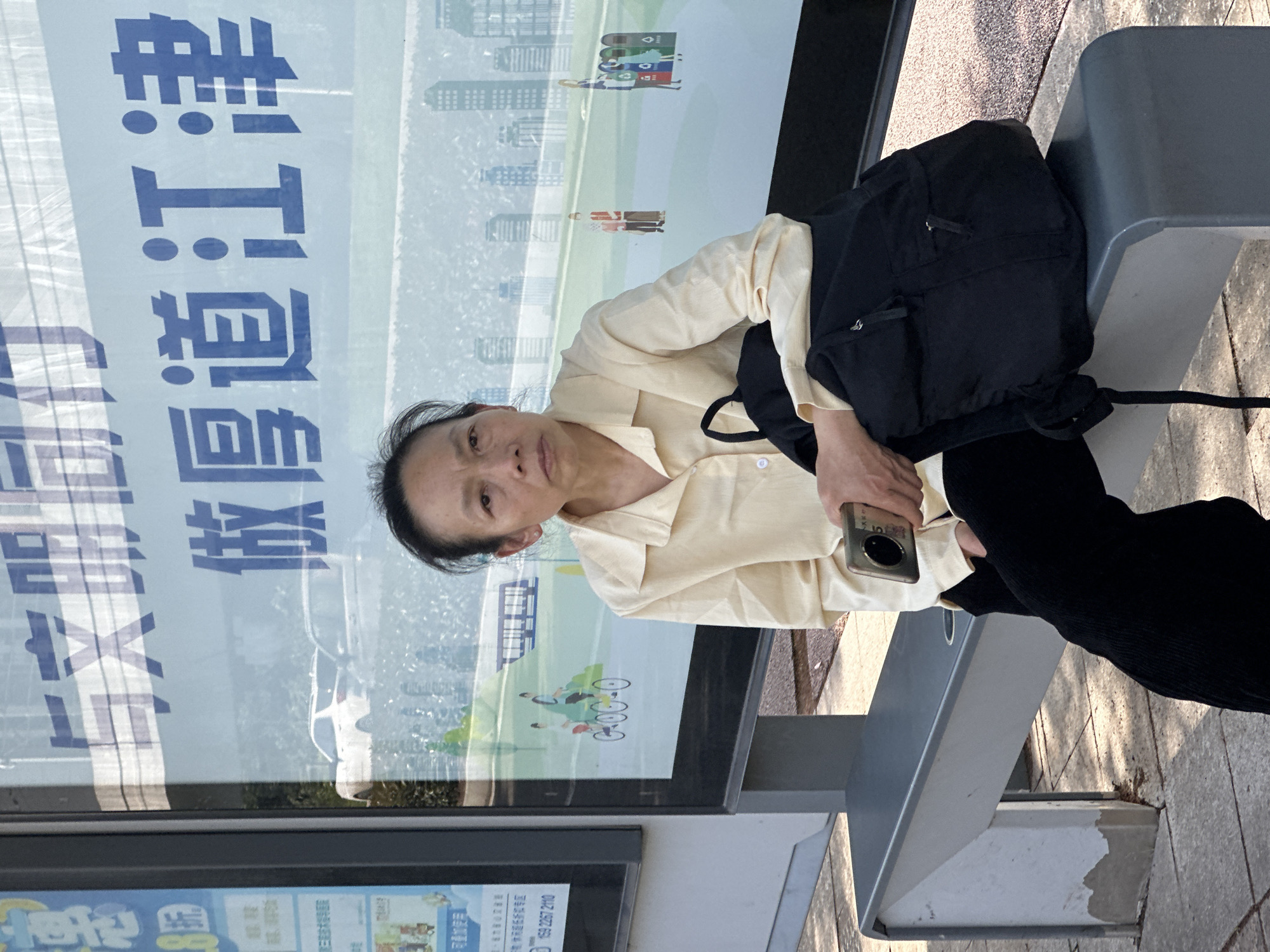 An adult, likely female, with dark hair pulled back, is seated on a grey public bench, looking directly at the viewer. She wears a light-colored collared shirt and dark pants, holding a smartphone in her right hand. A black backpack rests beside her on the bench. Behind her is a large display panel, possibly part of a bus stop or information board, featuring stylized illustrations of a city skyline, a bus, people walking and cycling, and recycling bins. The display also shows Chinese text which includes "做厚道江津" (Be honest/sincere, Jiangjin), "8號" (Number 8), "折" (Discount), "可叠加使用" (Can be used cumulatively), and a phone number "159 2267 2110". The scene appears to be an outdoor public setting during daytime, with clear weather.