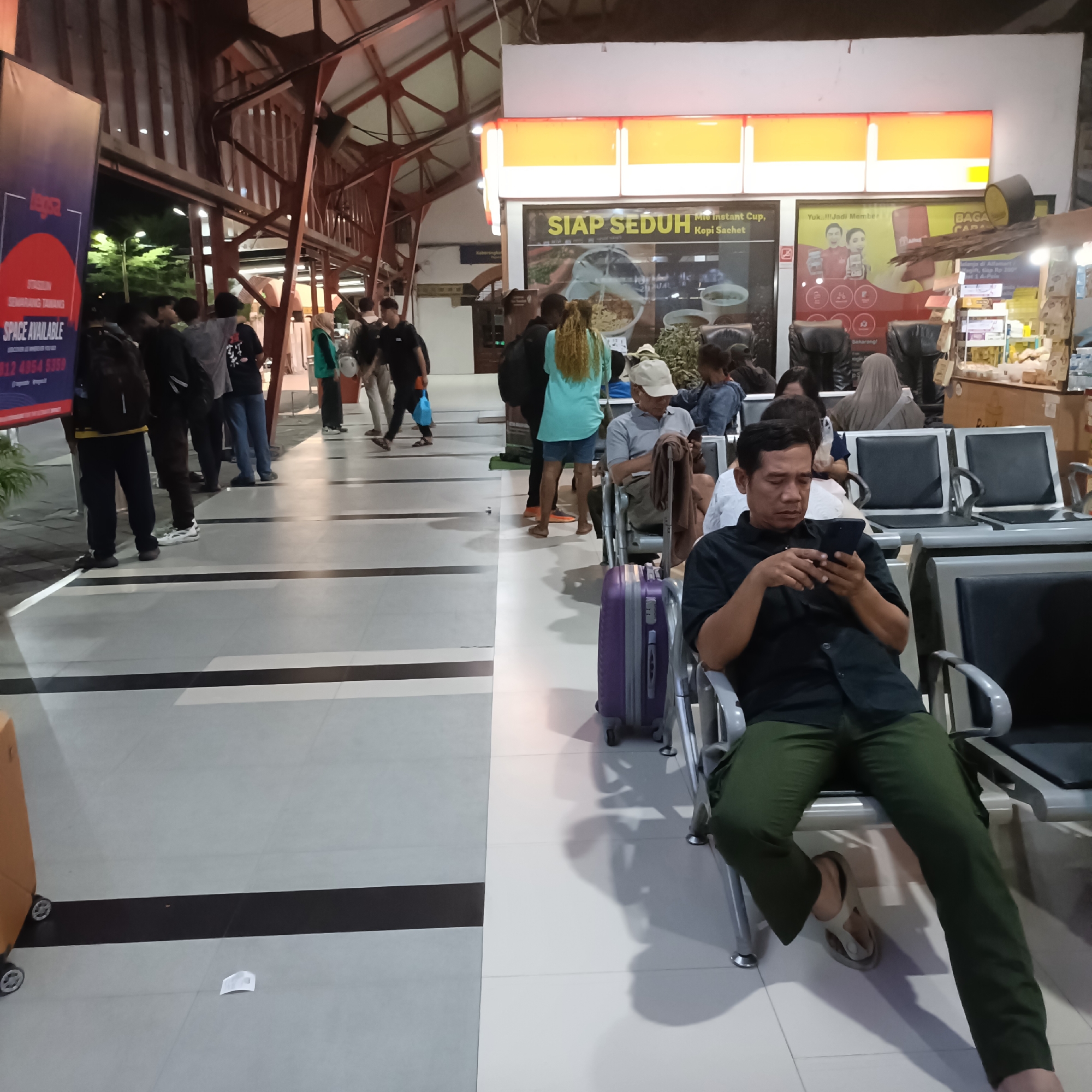 The image depicts a waiting area, possibly at a train station or bus terminal in Semarang, Indonesia. The setting appears to be indoors with a tiled floor. A prominent sign with the text "SIAP SEDUH" and an image of instant noodles is visible on the right side of the image, suggesting a food or convenience stall.
Several people are present, some standing and others seated in rows of chairs. In the foreground on the right, a man in a black shirt and green pants is sitting and looking at his phone. Beside him is a purple suitcase. Further back, other individuals are seen standing in groups or individually, some appearing to be waiting for transportation. A large banner with the text "SPACE AVAILABLE" is visible on the far left. The lighting suggests it is either evening or nighttime. The overall atmosphere appears to be that of a public transit waiting area.