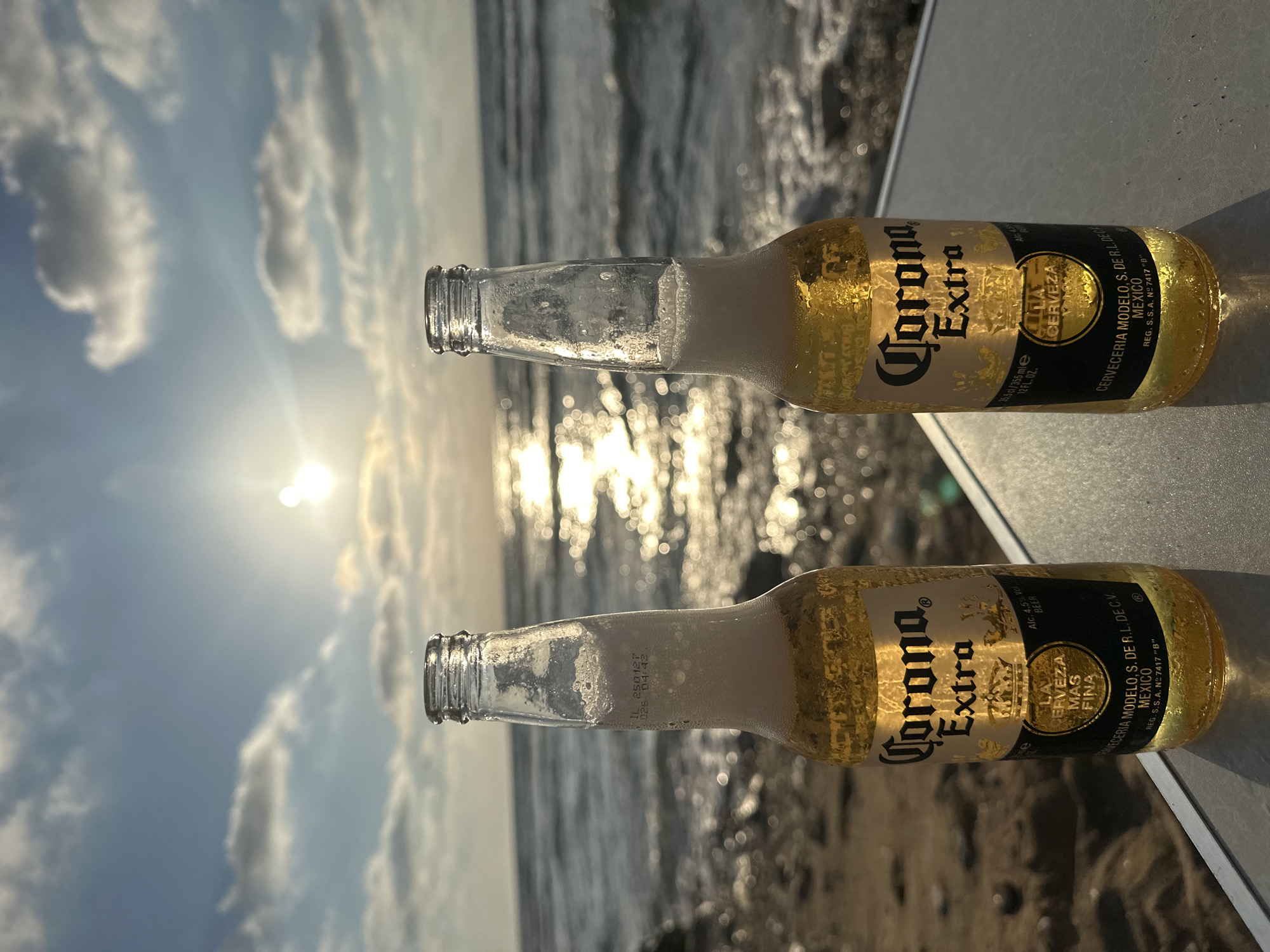 Two bottles of Corona Extra beer are placed on a table or surface in front of a beach with the sea and a sunset. The sky is blue with scattered white clouds, and the sun is setting over the water, casting a golden reflection. The bottles are partially filled with beer and have condensation on them. The labels on the bottles are clearly visible. In the background, there is a sandy beach with some rocks. The scene suggests a relaxed setting by the sea. The location is the city of Haifa, Israel.