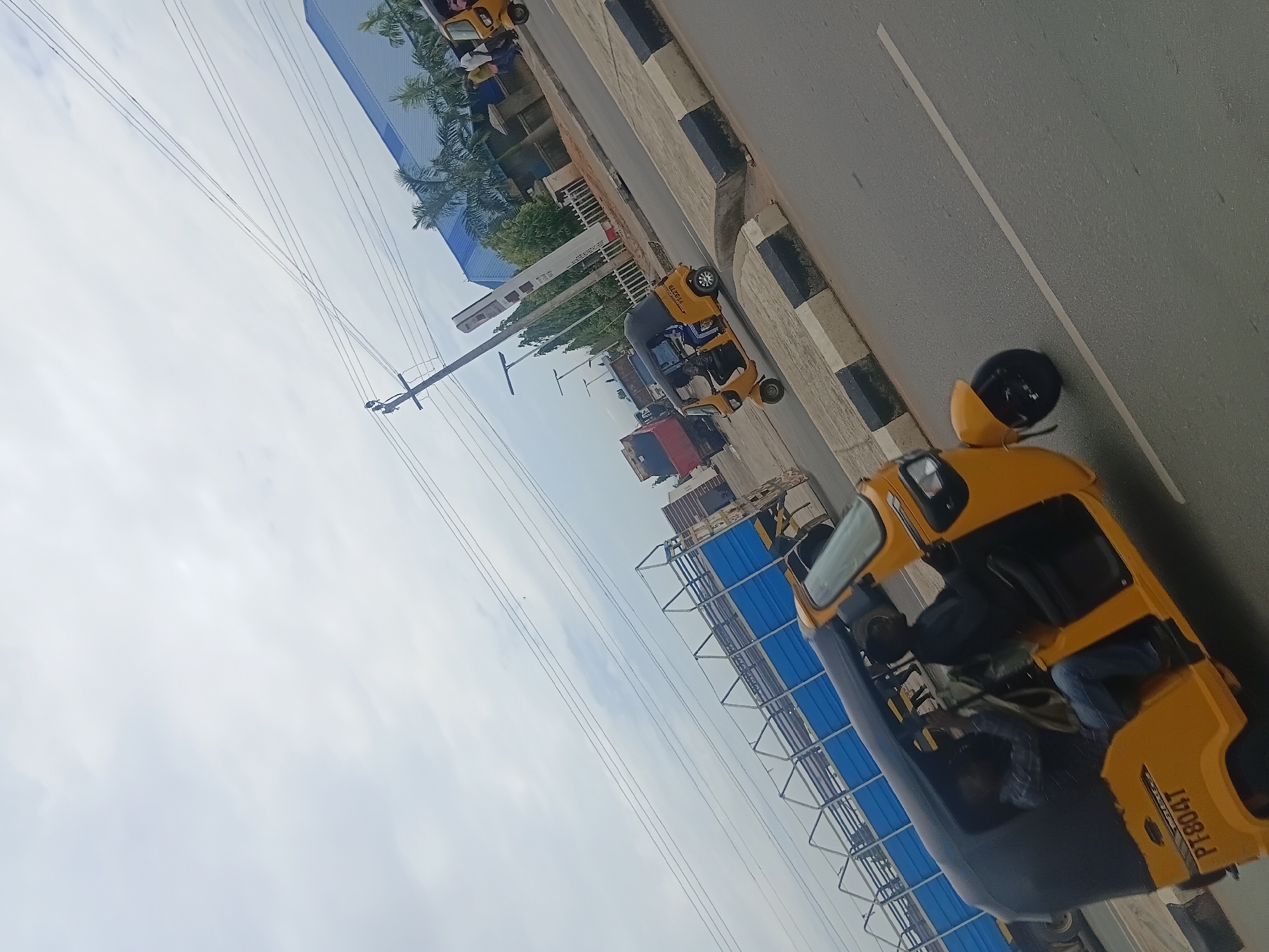 The image depicts a street scene in Jalingo, Nigeria, featuring several yellow auto-rickshaws, commonly known as "keke napeps" or "okadas" in the region. The vehicles are parked along a paved road, with a curb and sidewalk visible. In the background, there are buildings, some with blue roofing, and palm trees suggesting a tropical or subtropical climate. Power lines crisscross the sky, indicating an urban or semi-urban setting.
The time of day appears to be daytime, with the sky overcast, suggesting a cloudy or possibly humid weather condition. The auto-rickshaws are the primary objects, some with passengers or drivers visible inside. One auto-rickshaw has the text "PT8041" clearly visible on its rear. The overall impression is a bustling roadside environment, typical of many Nigerian cities. The focus of the image is on the transportation infrastructure and the vehicles that are integral to daily life and commerce in such locations.