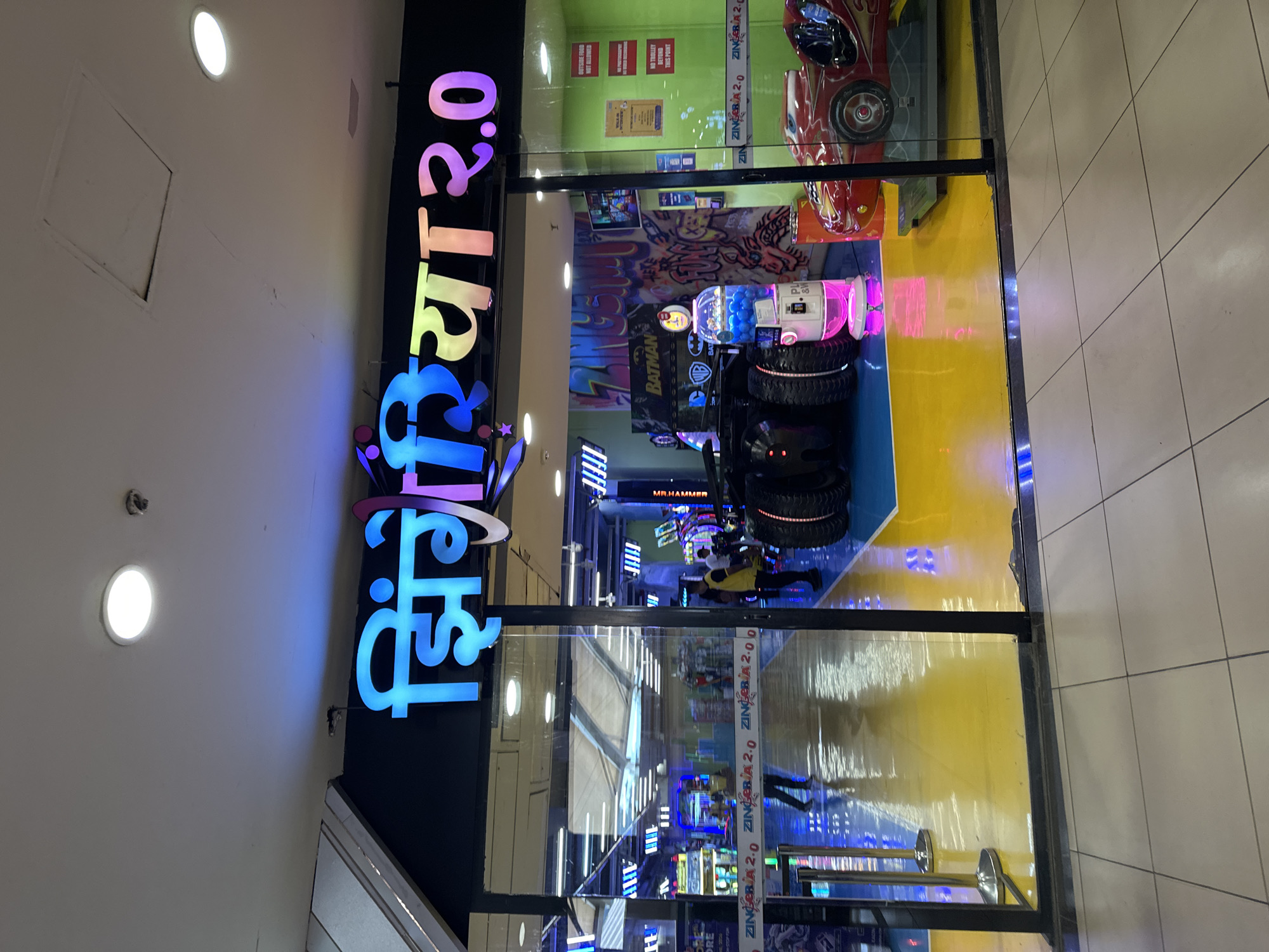 The image shows the entrance to "Zingeria 2.0", an arcade or entertainment center. The sign for Zingeria 2.0, with multicolored lettering, is prominent over the entrance. Inside, there are arcade games, including a Batman-themed game, a car racing game, and other machines with flashing lights. The floor is a combination of yellow and blue. The reflection shows the tile flooring. One or two people are visible inside the arcade. The location is likely in Kalyan-Dombivli, India.