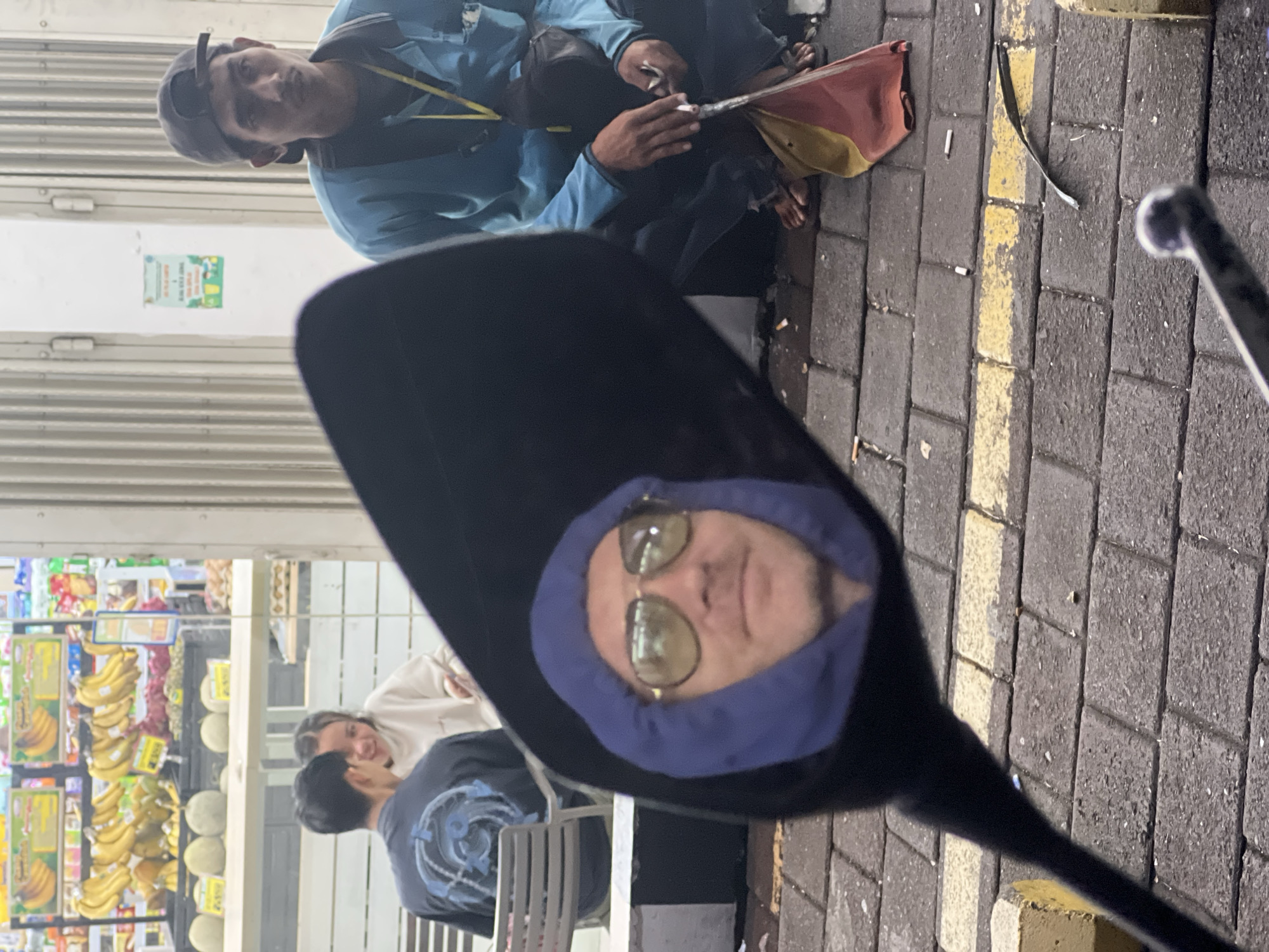 The image shows a reflection in a motorcycle mirror of a person with sunglasses and a blue hooded sweatshirt, sitting at a table with two other individuals; in the background, a fruit stand stocked with bananas and melons. In the foreground, blurred, is a man in a blue jacket and cap sitting on the ground, using a cigarette holder. The street is paved with grey bricks and has a yellow line on the side. Based on the fruit and the man with the cigarette, it appears to be a roadside scene in Denpasar, Indonesia.