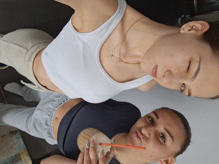 The image displays two young females in an indoor setting within Sofia, Bulgaria.
The female on the left, positioned closer to the camera and angled slightly, has light brown hair, fair skin, and blue eyes. She is wearing a light grey ribbed crop top and light beige ribbed sweatpants with a visible drawstring. Her right eye is winking, and her lips are slightly pursed. She has multiple piercings on her left ear, including a hoop, a small stud, and a small cross-shaped stud. Two layered gold necklaces are visible, one featuring small circular charms and a horseshoe pendant, and the other a small cross pendant.
The female on the right has dark hair pulled back and a medium skin tone. She is wearing a dark grey or navy blue cropped t-shirt and light grey sweatpants, with the word "FAMILI" partially visible on the waistband. She holds a clear plastic cup containing a light brown beverage with a white topping (possibly whipped cream) and an orange straw. Her left hand, visible on the cup, has styled fingernails. Her lips are pursed in a pout, and she is looking towards the camera.
The background consists of a plain light grey wall on the right and a dark, indistinct surface or furniture item on the left. A small portion of what appears to be a multi-colored rug or abstract artwork is visible in the lower right corner. The lighting is consistent with an indoor environment. No distinct exterior landmarks or specific interior identifiers are present to pinpoint an exact address or known public venue within Sofia.