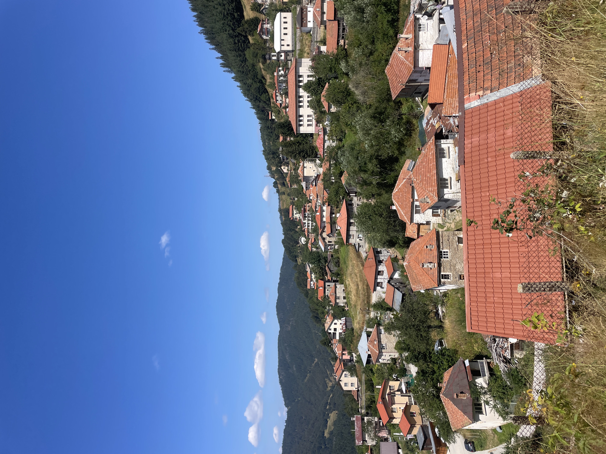 The image shows a panoramic view of a hillside village in Manastir, Bulgaria, under a clear blue sky with a few scattered clouds. The village is nestled amongst rolling green hills covered with dense forests of pine trees. The architecture consists of numerous houses, many with characteristic red tile roofs, built in a traditional style. The houses are of varying sizes and colors, with some appearing to be made of stone and others of stucco.
The landscape is rugged, with the houses built on different levels of the slope, creating a charming and somewhat chaotic urban fabric. Vegetation is abundant, with trees and shrubs interspersed among the buildings and covering the hillsides. In the foreground, a chain-link fence with some dry grass and weeds can be seen, suggesting a slightly overgrown or untamed area.
Several cars are visible parked on a road winding through the village, indicating that it is an inhabited community. One prominent building with a white facade and a dark steeple stands out, possibly a church or a public building. The overall impression is of a peaceful, rural setting in a mountainous region. There are no people visible in the image.