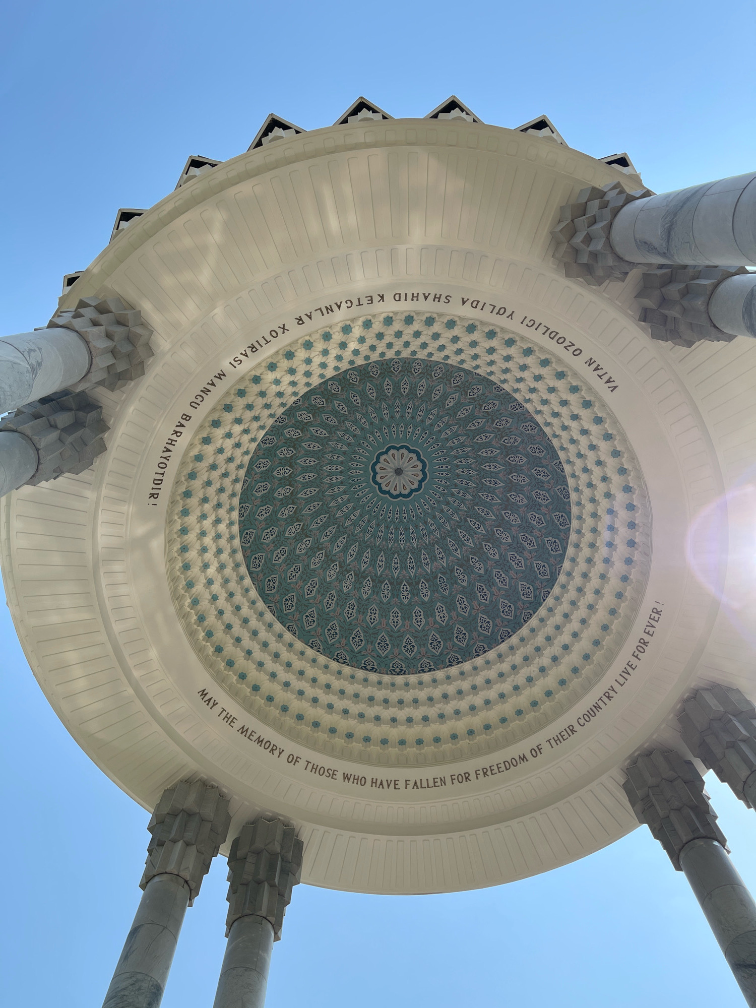 The image shows a low-angle view of the dome of the Independence Monument in Tashkent, Uzbekistan. The monument is a large, white structure with intricate carvings and a blue and white patterned dome. The dome is supported by four large, marble columns. The sky above is clear and blue. There are inscriptions on the dome. One inscription reads: "MAY THE MEMORY OF THOSE WHO HAVE FALLEN FOR FREEDOM OF THEIR COUNTRY LIVE FOR EVER!" Another inscription, in Uzbek, is also visible. The sun is shining, creating a lens flare on the right side of the image. The monument appears to be empty, with no people visible.