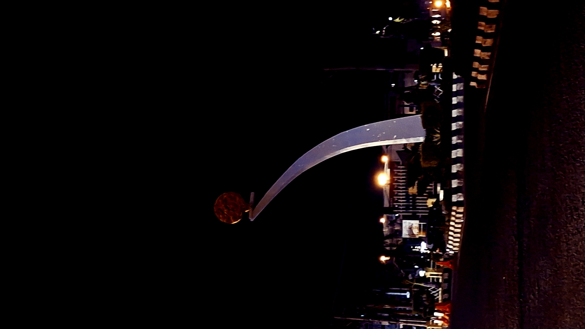 The image shows a monument at night in Lhokseumawe, Indonesia. The monument features a curved, white structure topped with a circular, textured object, possibly representing the sun. The scene is illuminated by streetlights and lights from nearby buildings, casting a warm glow on the surroundings. The foreground consists of a road with striped barriers and traffic cones, indicating it is likely a public space or intersection. The sky is dark, emphasizing the illuminated monument and cityscape.