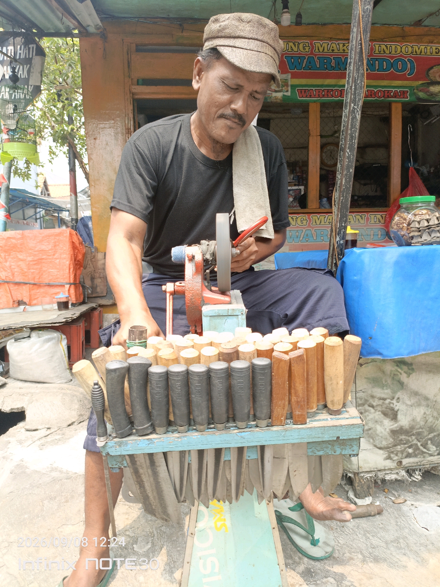 This is a medium shot, eye-level, outdoor photograph of a man sharpening knives in Central Jakarta, Indonesia. The man, wearing a black t-shirt and a brown cap, is seated in front of a sharpening machine with a red body. He is holding a red tool, possibly pliers, and appears to be working on a knife. In front of him is a wooden rack holding many knives with wooden and black handles. Behind him, a stall displays various food items. The background shows buildings and trees. The overall scene suggests a busy street market or roadside stall.