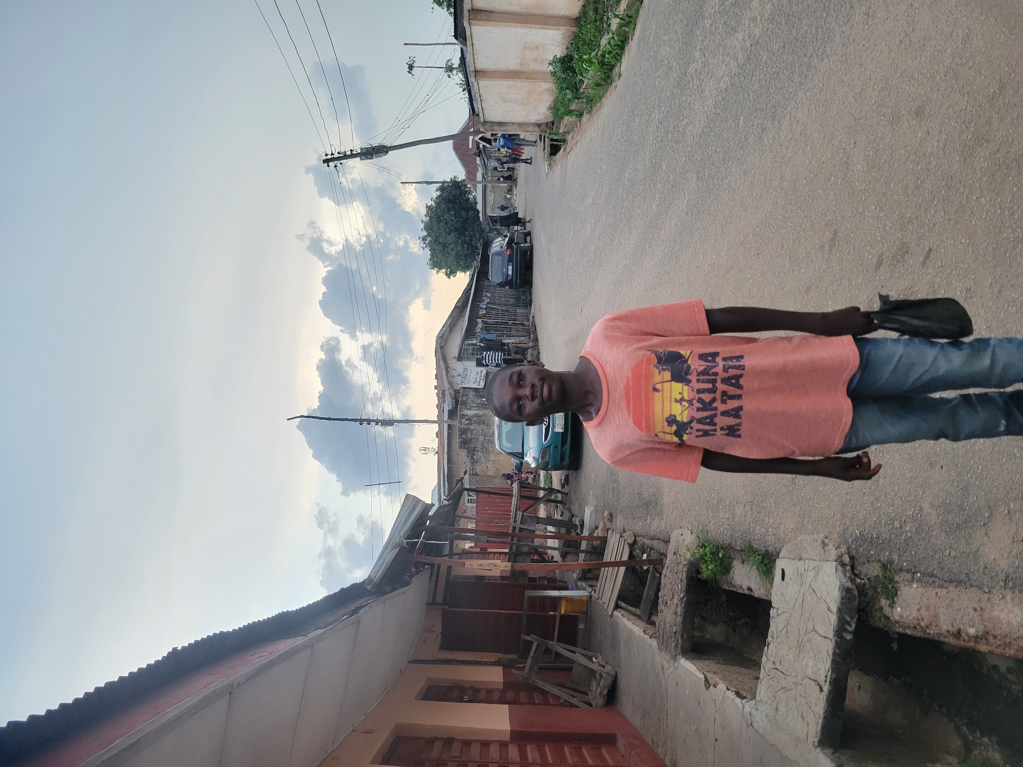 A person, likely a young male, stands on a paved street in what appears to be Jos, Nigeria. He is wearing a faded orange t-shirt with the words "Hakuna Matata" printed on it, along with blue jeans. He is holding a dark object in his hand.
The environment includes a street with buildings of varying construction styles and materials on either side. There is exposed drainage infrastructure running along one side of the street. Power lines are visible overhead. In the background, other people and vehicles are present on the street. The sky is overcast.