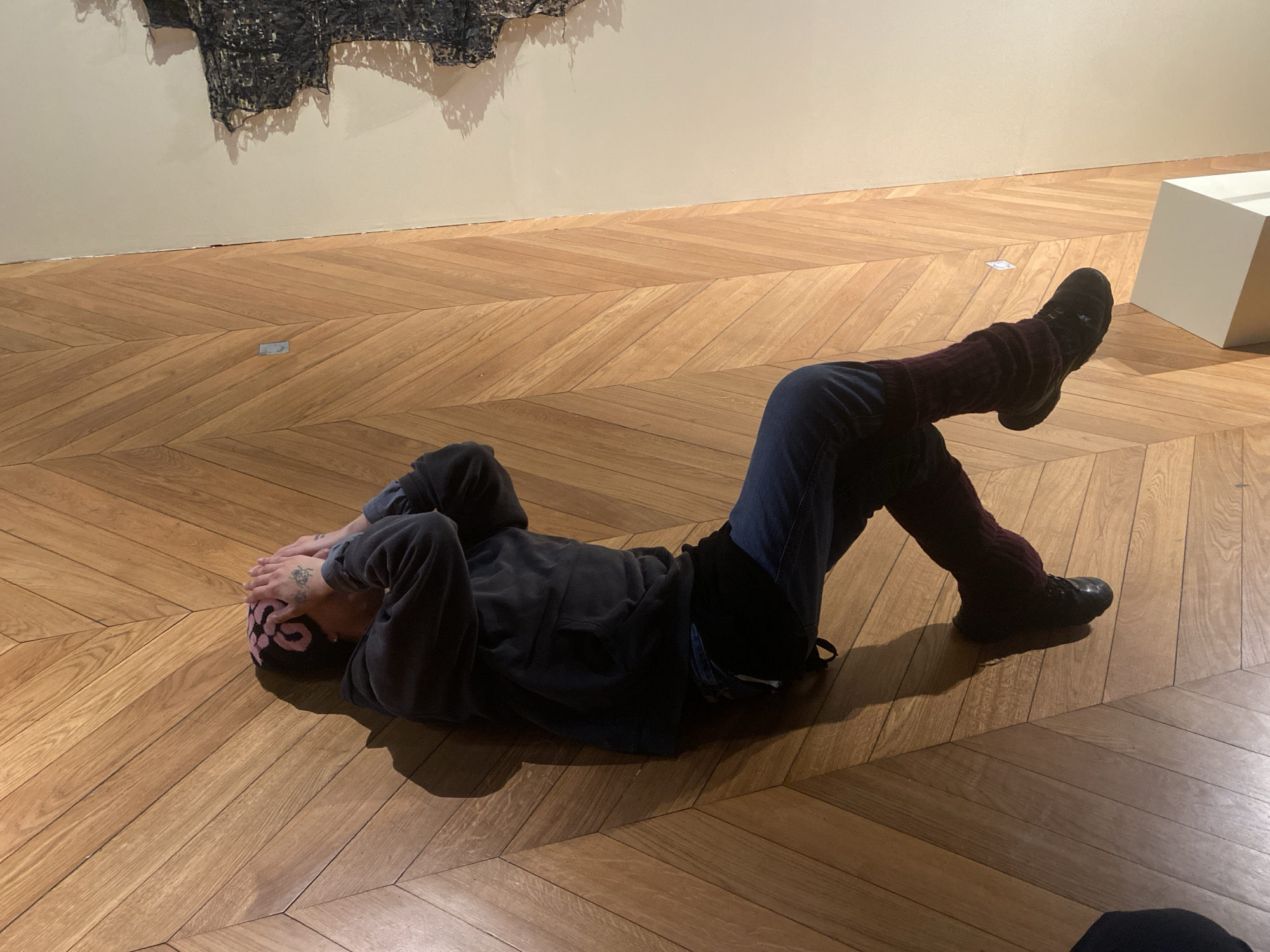 A person lies on their back on a parquet wood floor, with their head near a wall displaying an abstract artwork. The person is wearing a black beanie with pink designs, a dark grey sweatshirt, jeans, dark maroon leg warmers, and black shoes. They are positioned diagonally across the frame, with their head to the left and feet to the right, raised slightly off the floor. Their hands cover their face. In the background, a white rectangular prism sits on the floor. The floor is made of light-brown wooden planks arranged in a herringbone pattern. The location is likely an art gallery or museum in Paris, France, given the art on the wall and the architectural style.