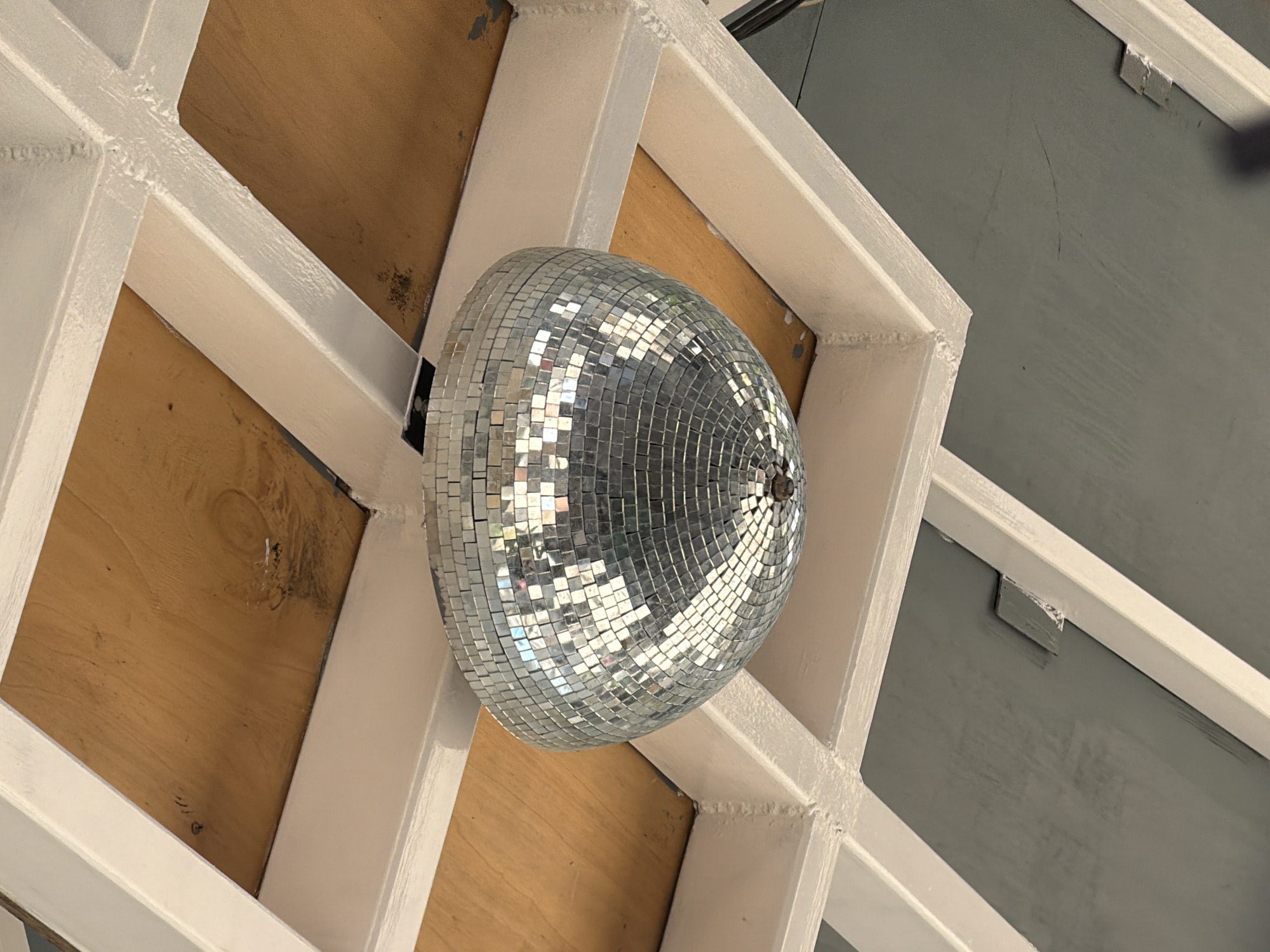 The image shows a disco ball hanging from a ceiling structure. The ceiling is made of white-painted beams forming a grid, with wooden panels filling some of the sections. The disco ball is spherical and covered in small mirrored squares, reflecting light. The overall scene appears to be indoors, possibly in a venue or recreational space, and does not contain any identifiable landmarks or features that would indicate a specific location in Varna, Bulgaria. There are no people or actions depicted in the image. The environment is static and focused on the disco ball and the ceiling.